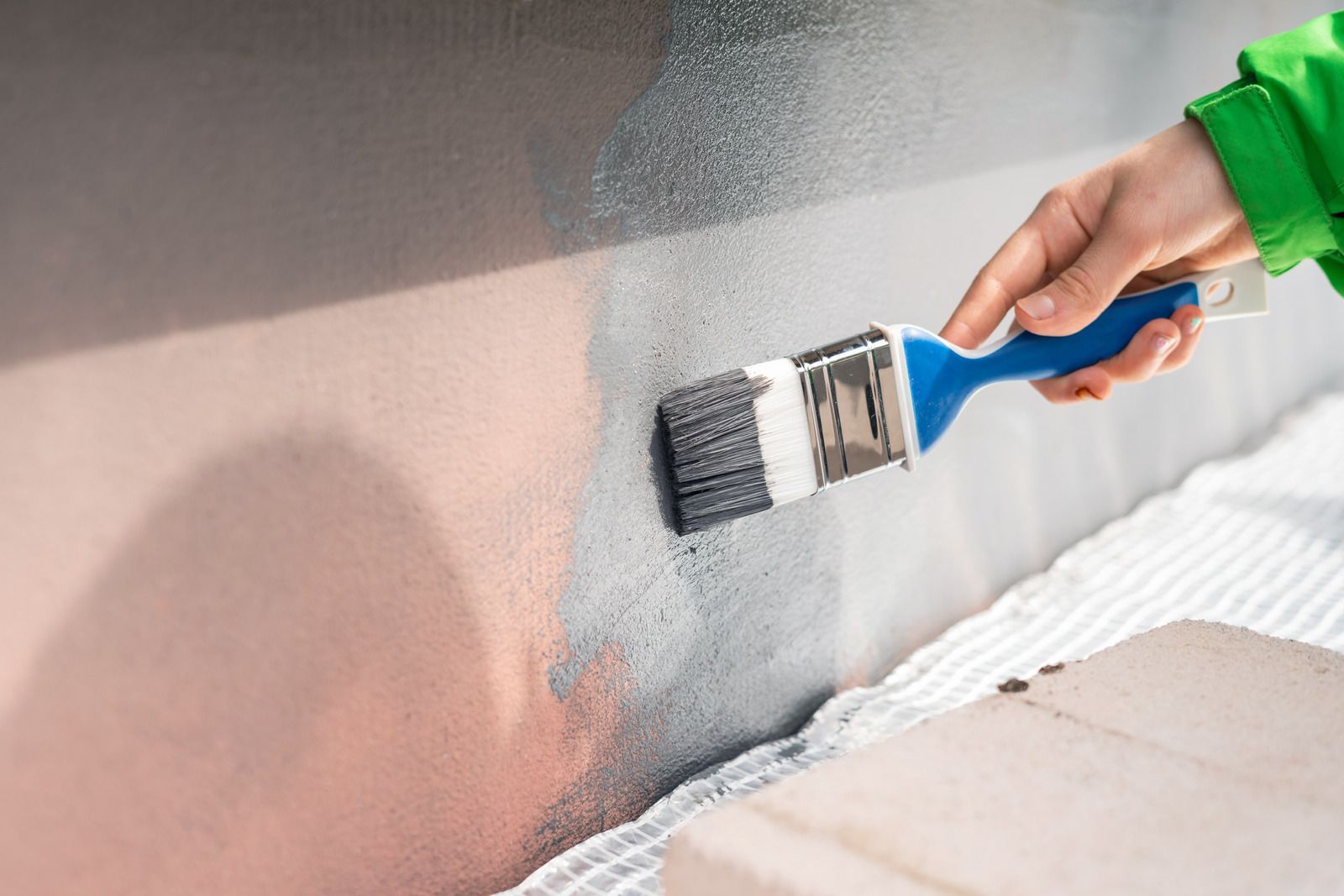 Hand painting a wall gray with a paintbrush near masking tape and drop cloth