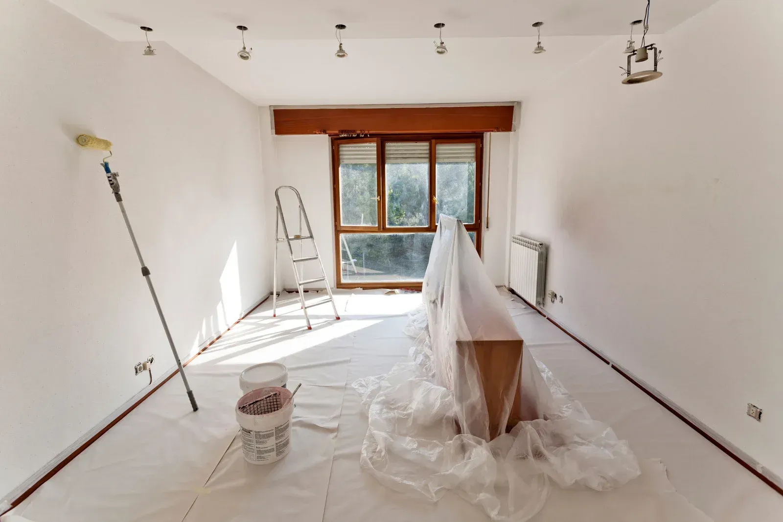 Room under renovation, walls painted white, ladder, roller, paint cans, furniture covered, window.