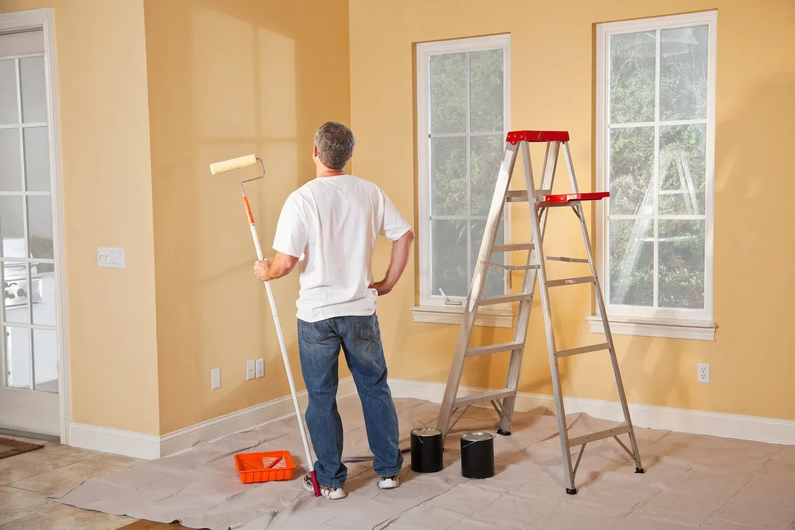 A person stands in a room with tan walls, holding a paint roller, with a metal ladder and paint cans nearby.