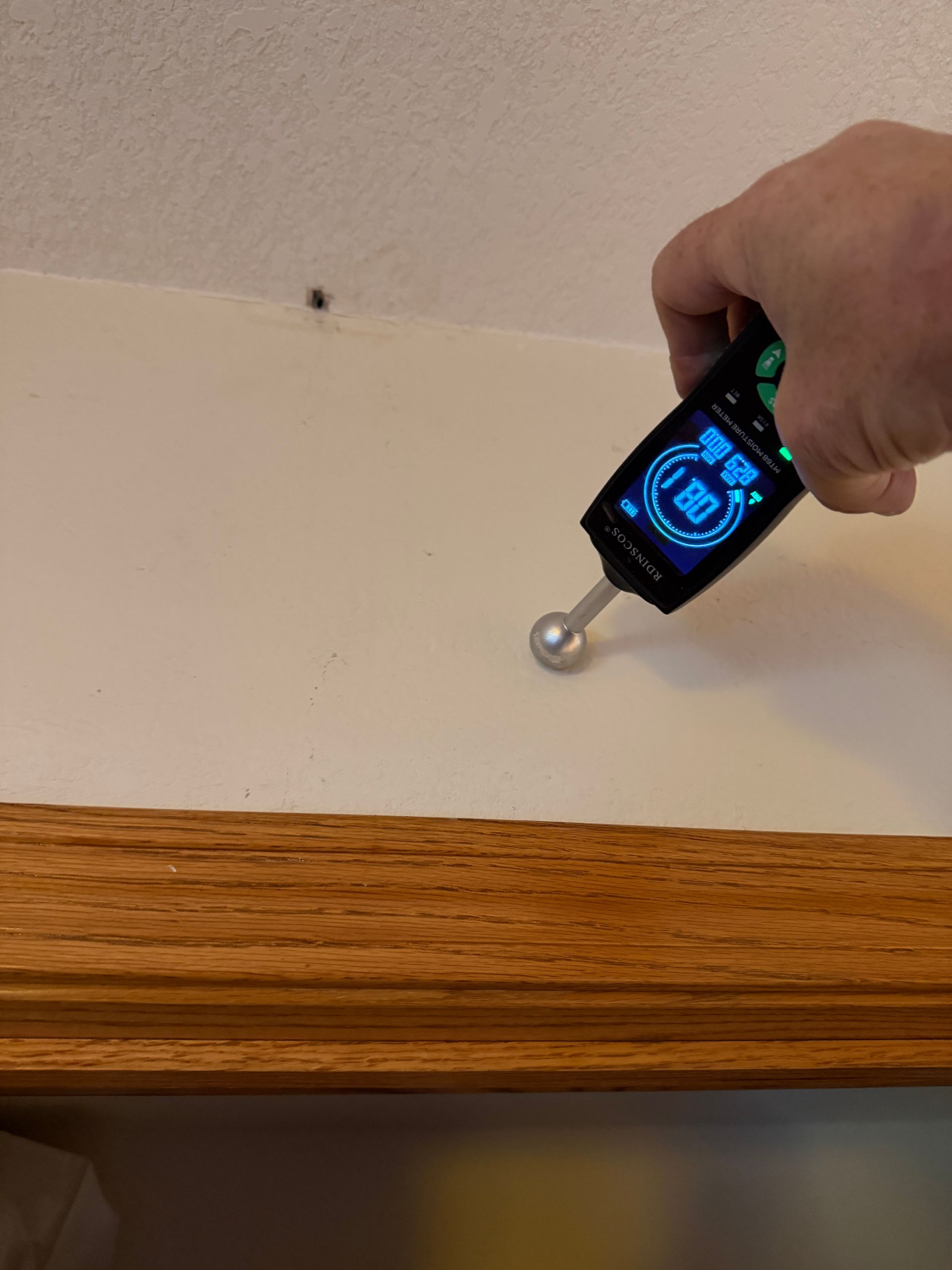 Hand holding moisture meter against a white wall near a wooden trim. The meter reads 83.