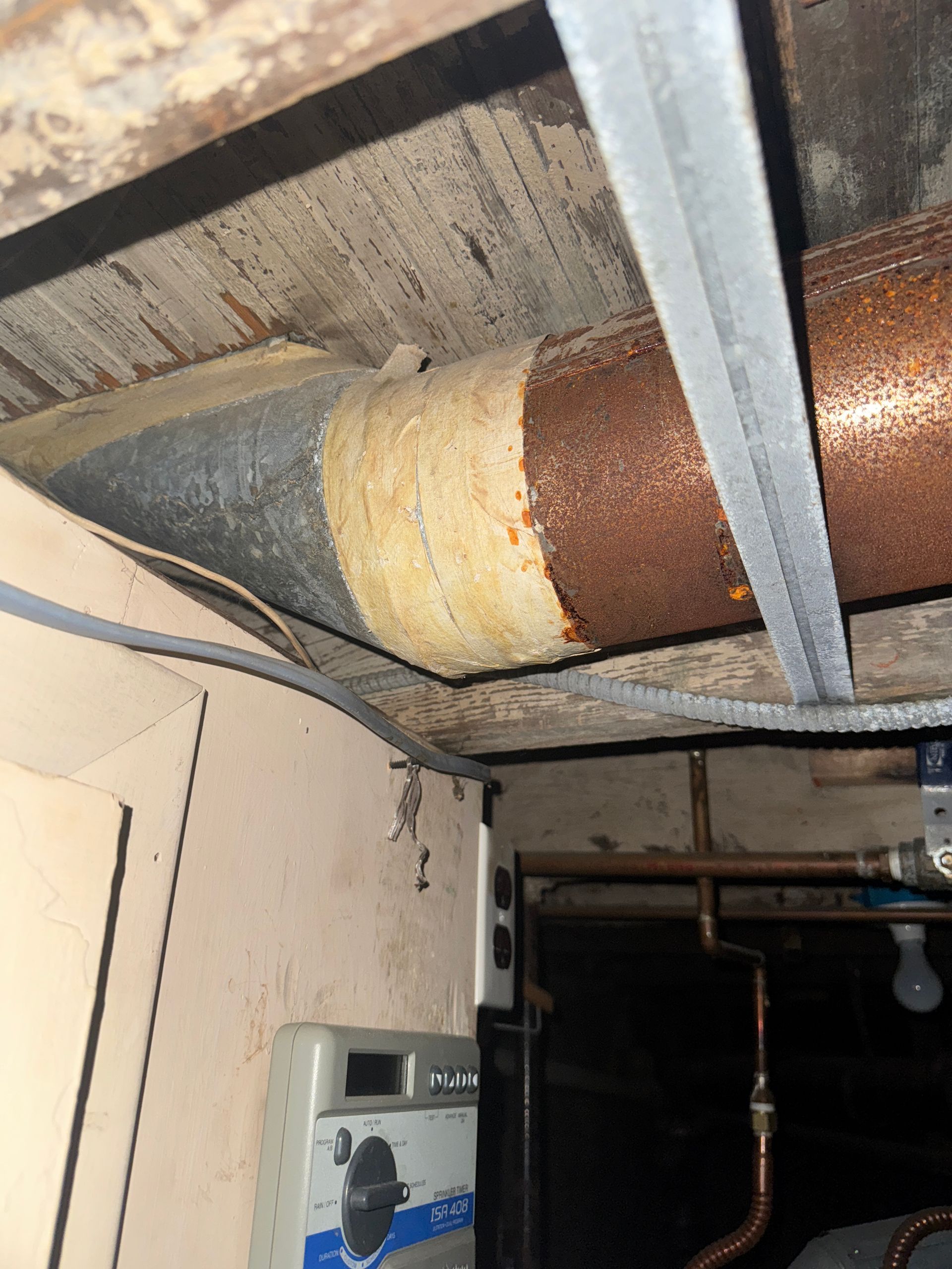 Rusty pipes and ductwork in a basement ceiling, with a white electrical box on the wall.