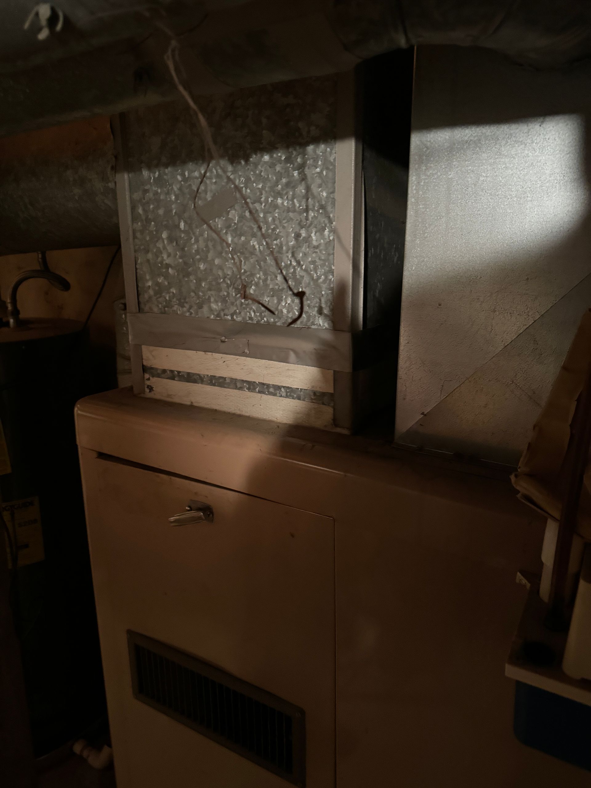 Furnace unit with filter, inside a dark basement. Silver metal ducts and filter box, beige furnace.