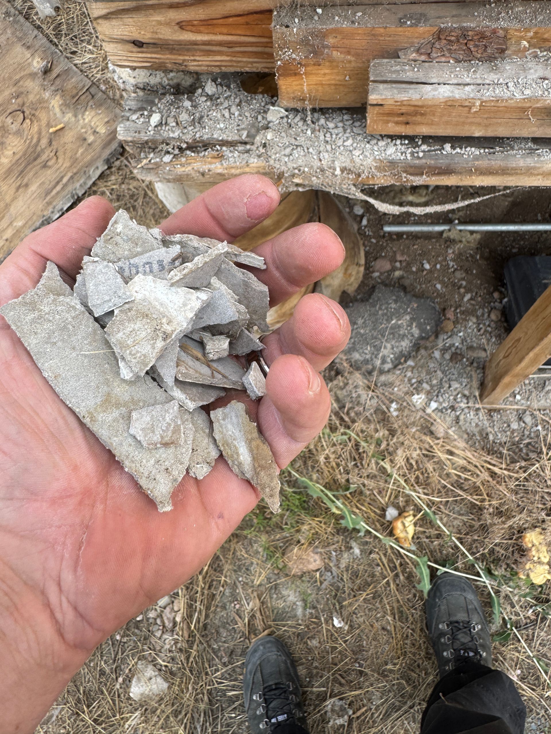 Hand holding pieces of light gray, flaky material; sawdust and wood in the background.
