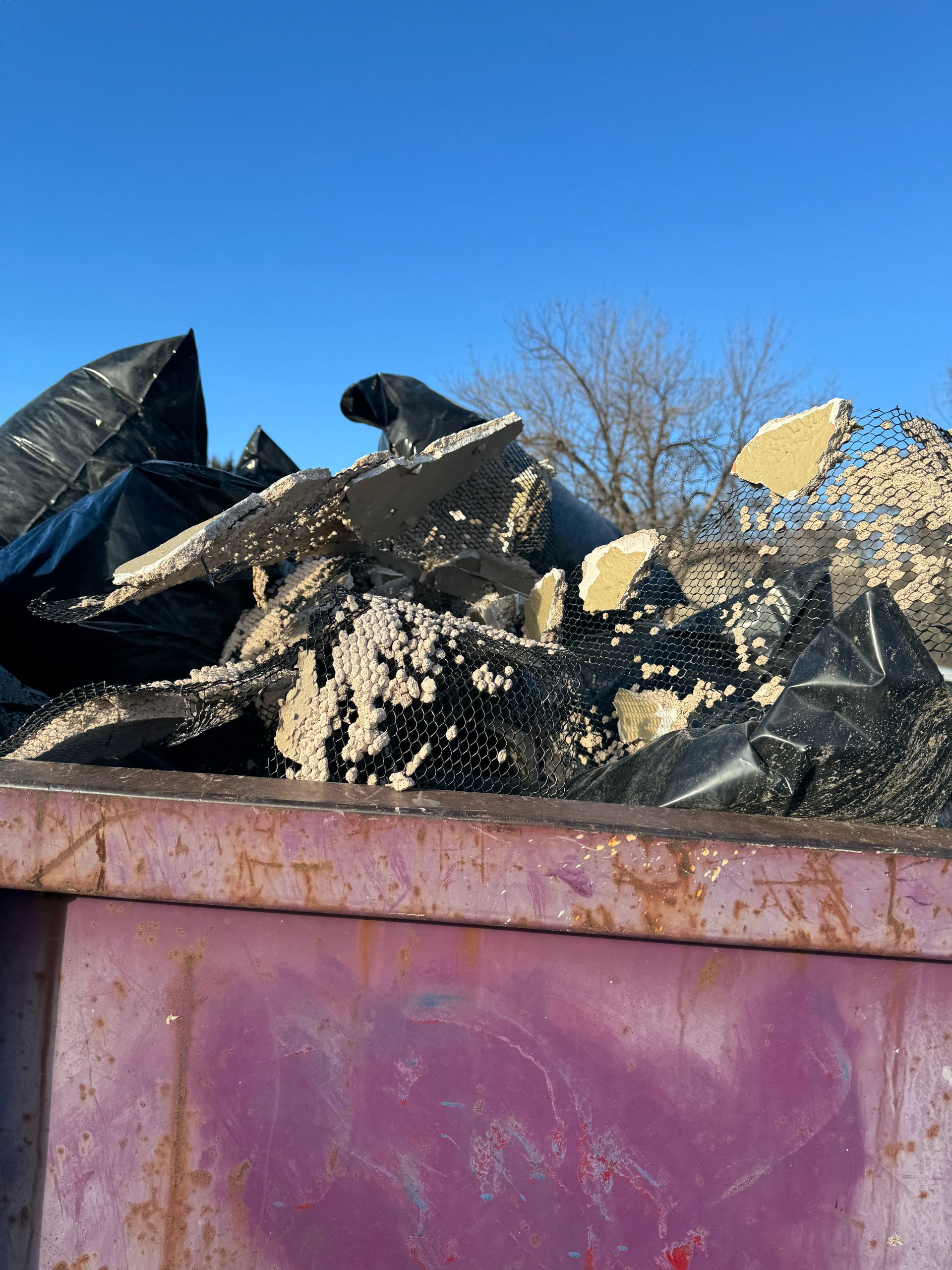 A rusty purple dumpster overflowing with broken drywall and black trash bags under a clear blue sky.