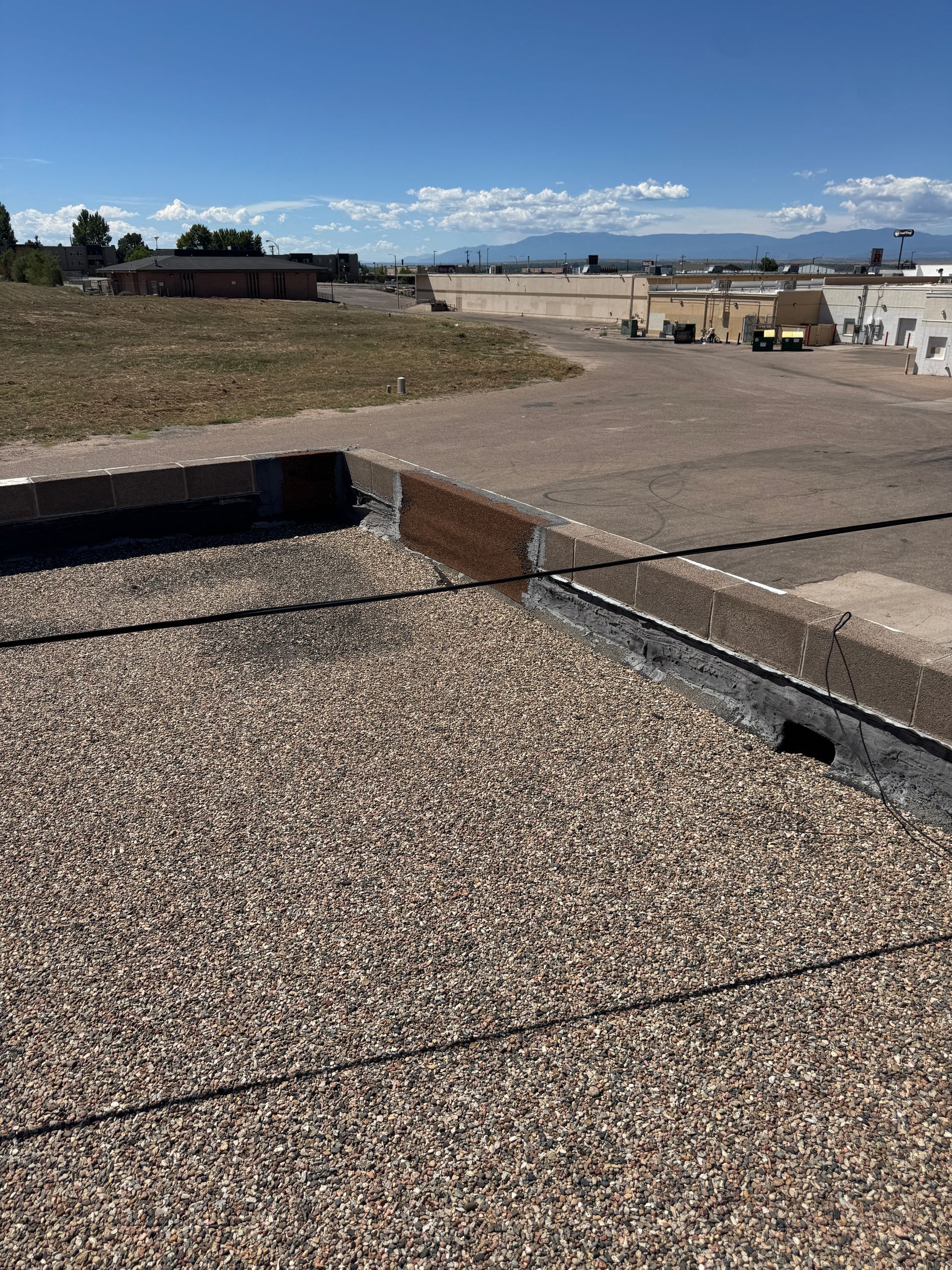 View from a gravel-covered rooftop, showing surrounding buildings and a patch of grass under a blue sky.