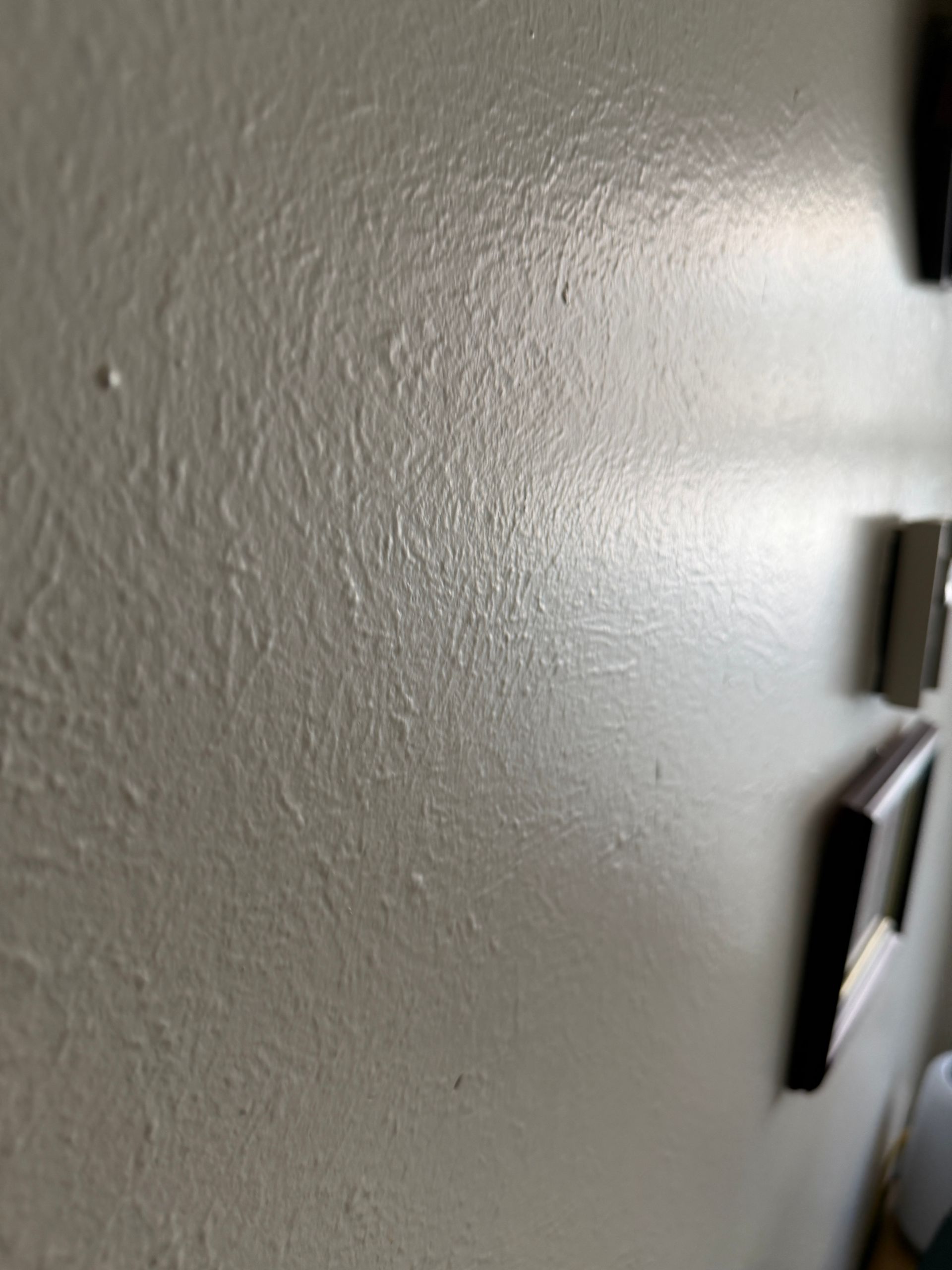 Textured, beige wall, angled view. Shadowed, with visible brushstrokes. Dark picture frames partially visible on right.