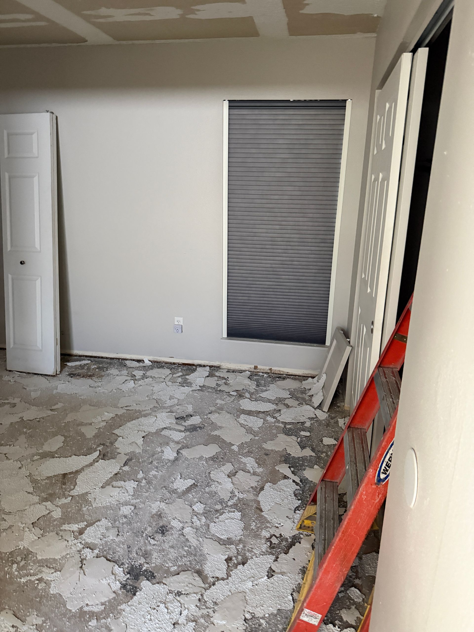 Room under renovation with debris on the floor, unfinished walls, closed blinds, and a leaning ladder.