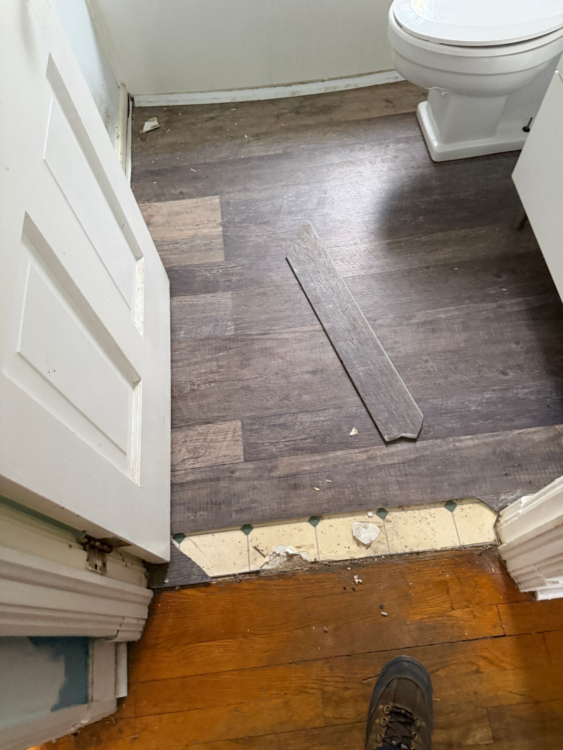 Bathroom floor with damaged flooring, wood floor transition and ajar white door.