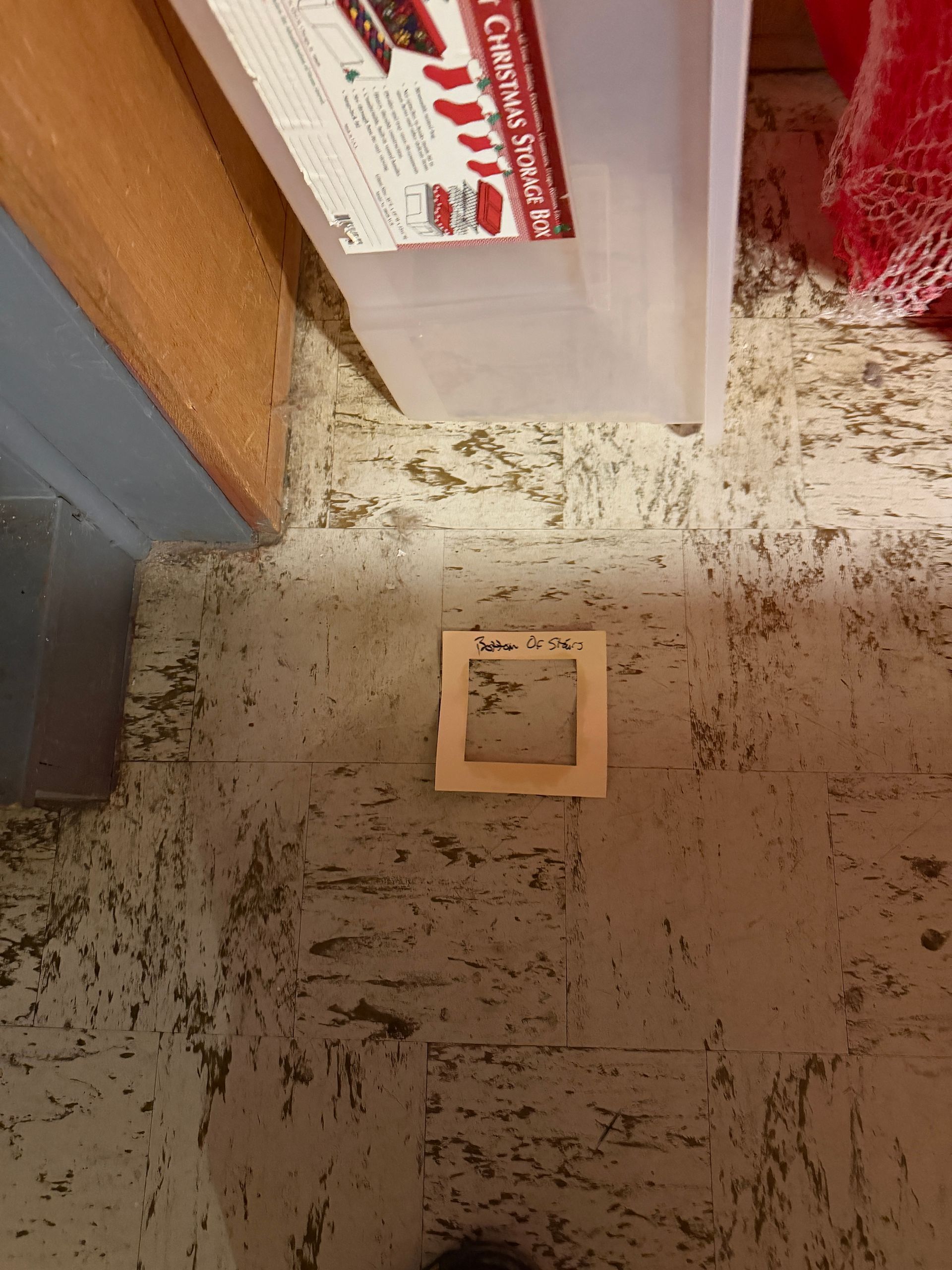 A square piece of paper with handwritten text sits on a worn, speckled floor, near a cabinet and a water jug.