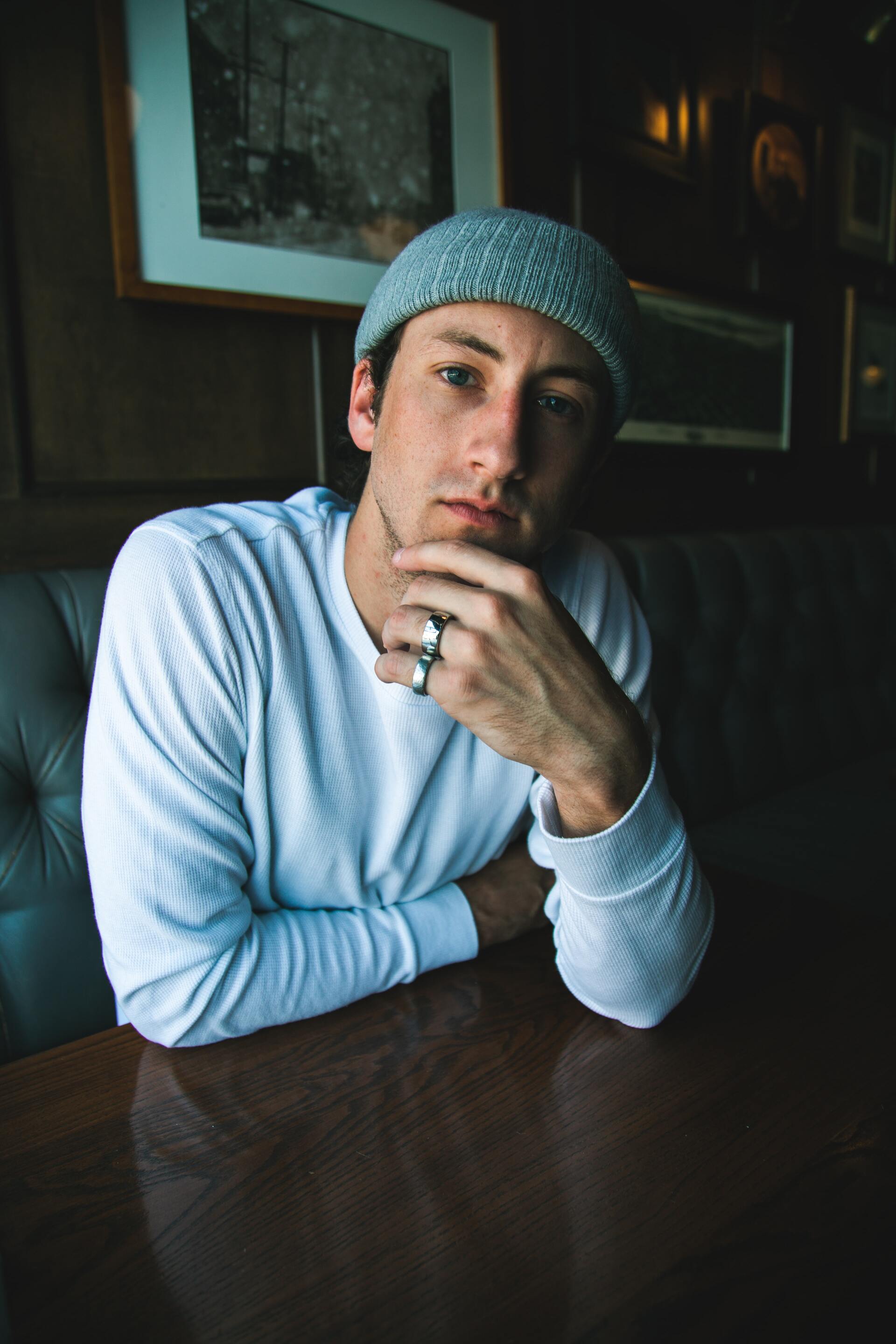 A man wearing a beanie and a ring is sitting at a table with his hand on his chin.