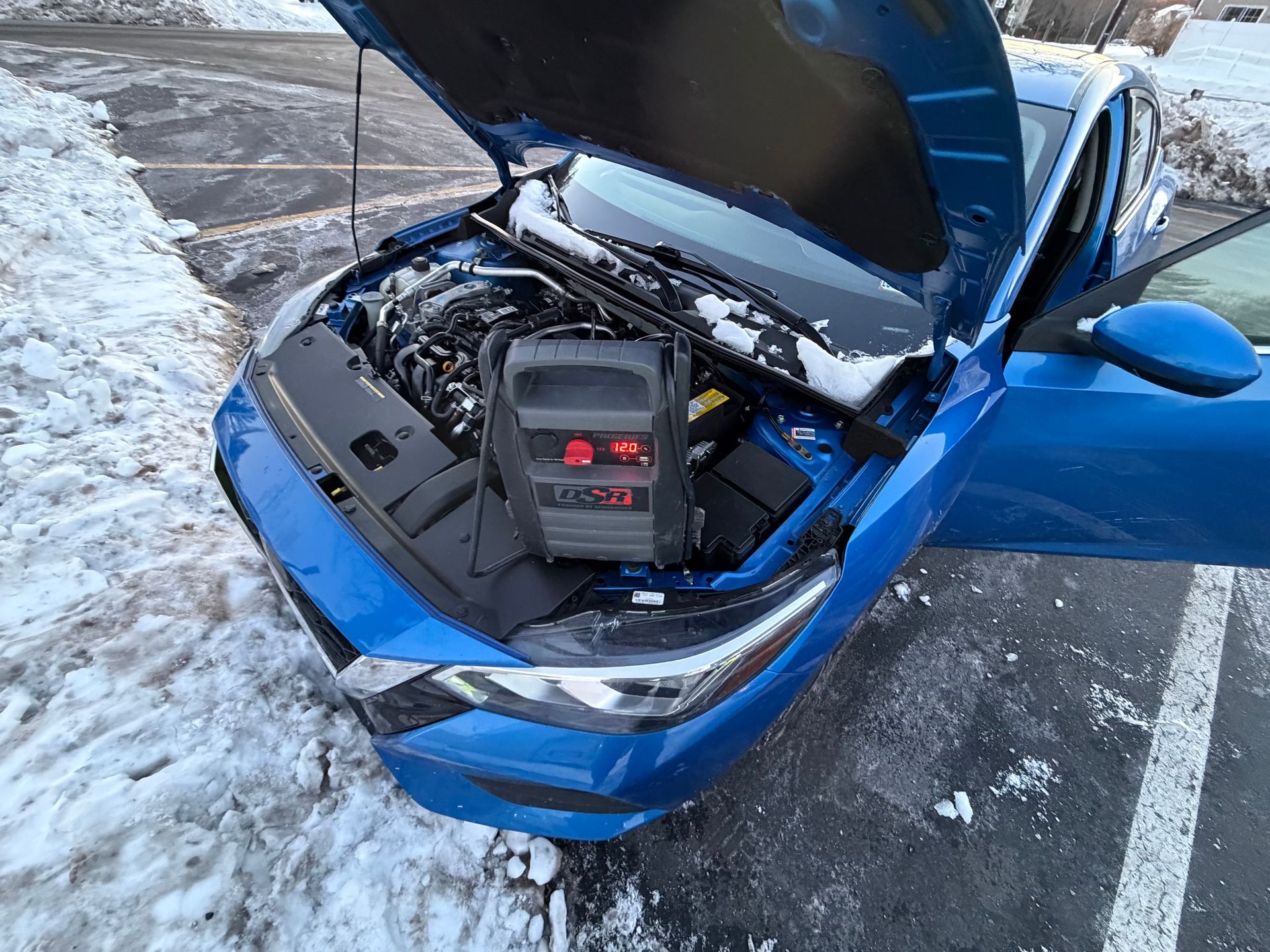 Blue car with open hood showing engine bay and battery in a snowy parking lot