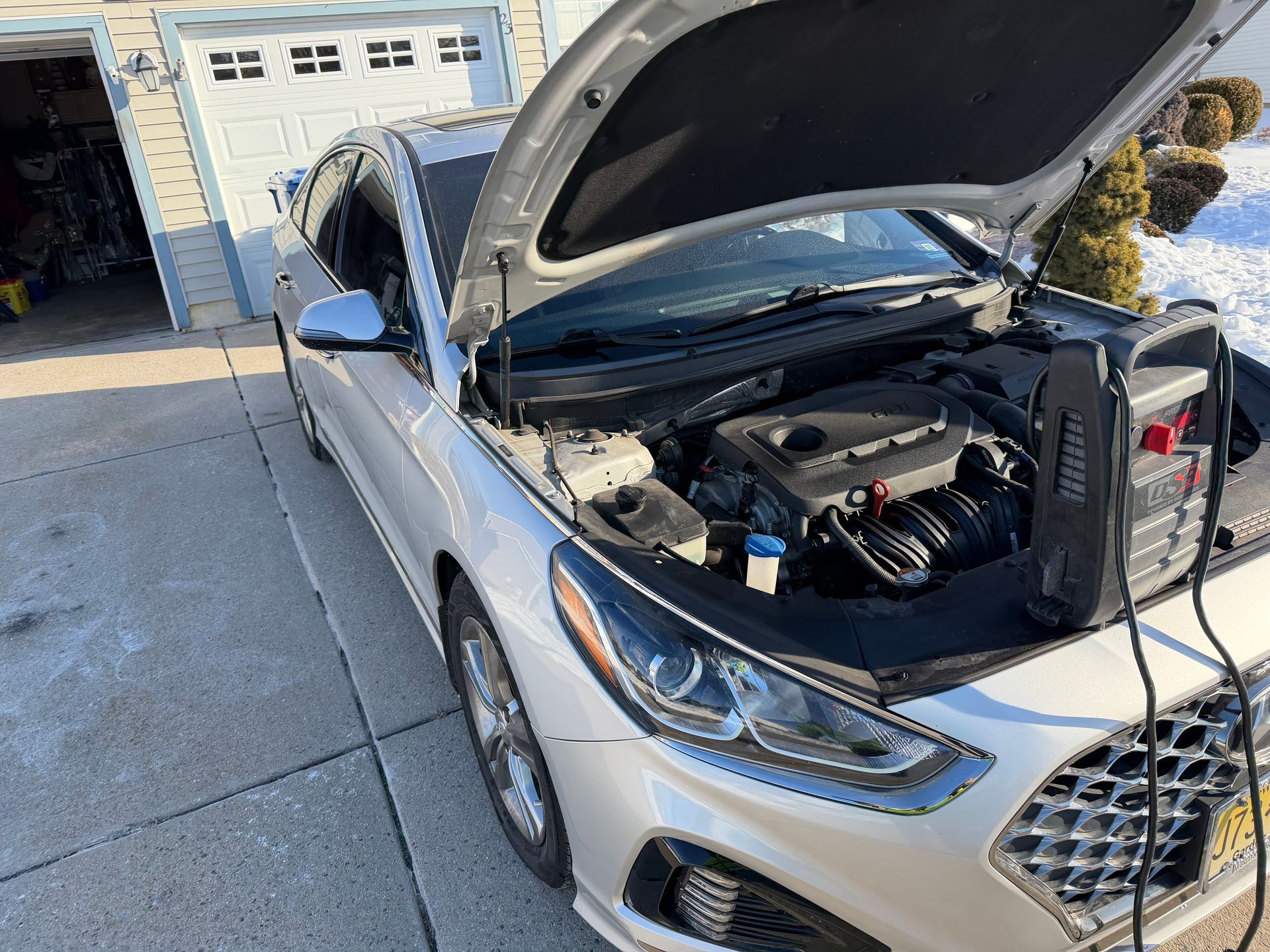 Silver car with open hood and battery charger connected in a driveway