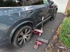 Dark SUV with a wheel clamp parked beside a driveway and brick wall, red tools on the ground