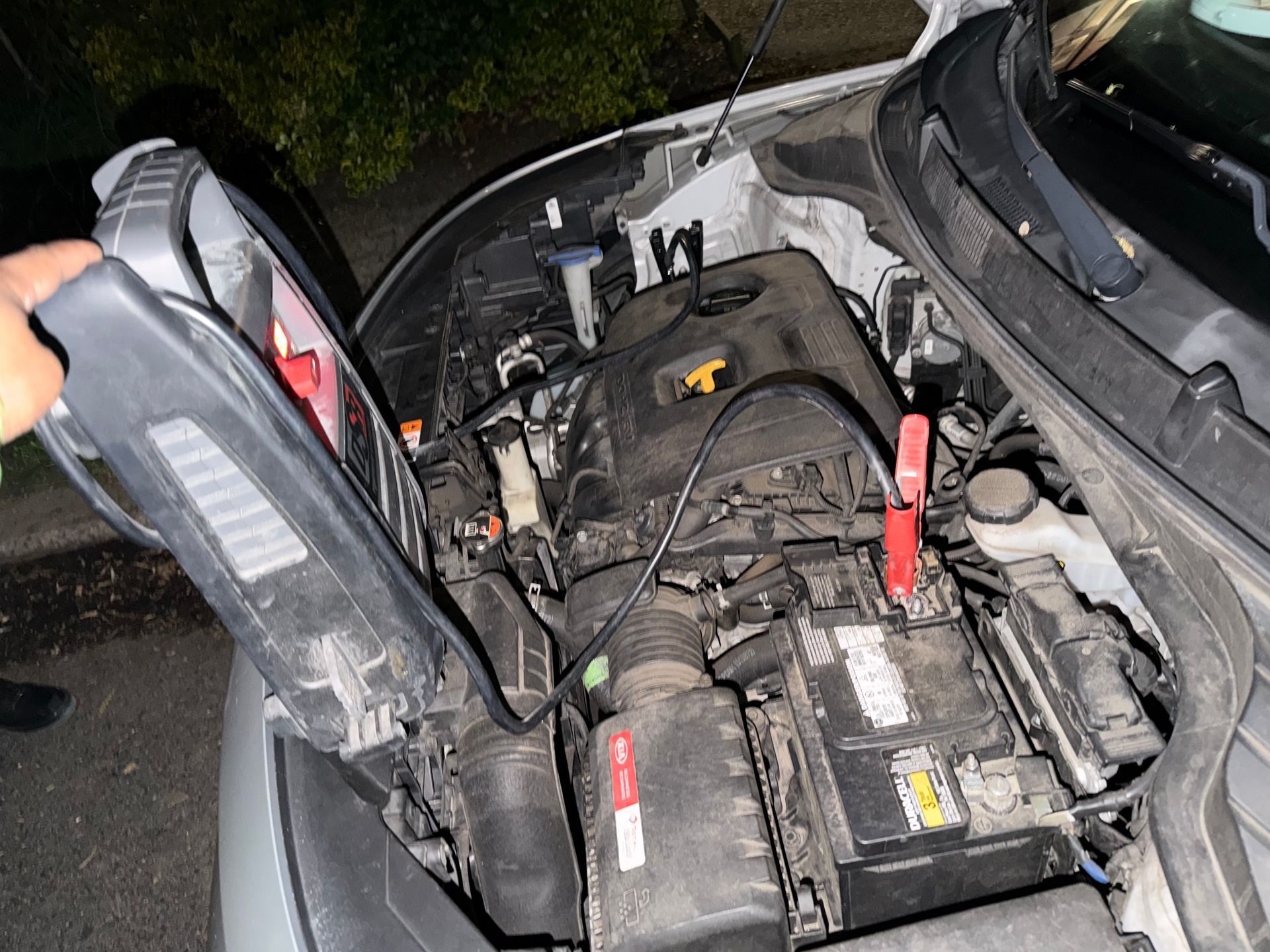 Open car hood showing engine bay with battery and jumper cables at night