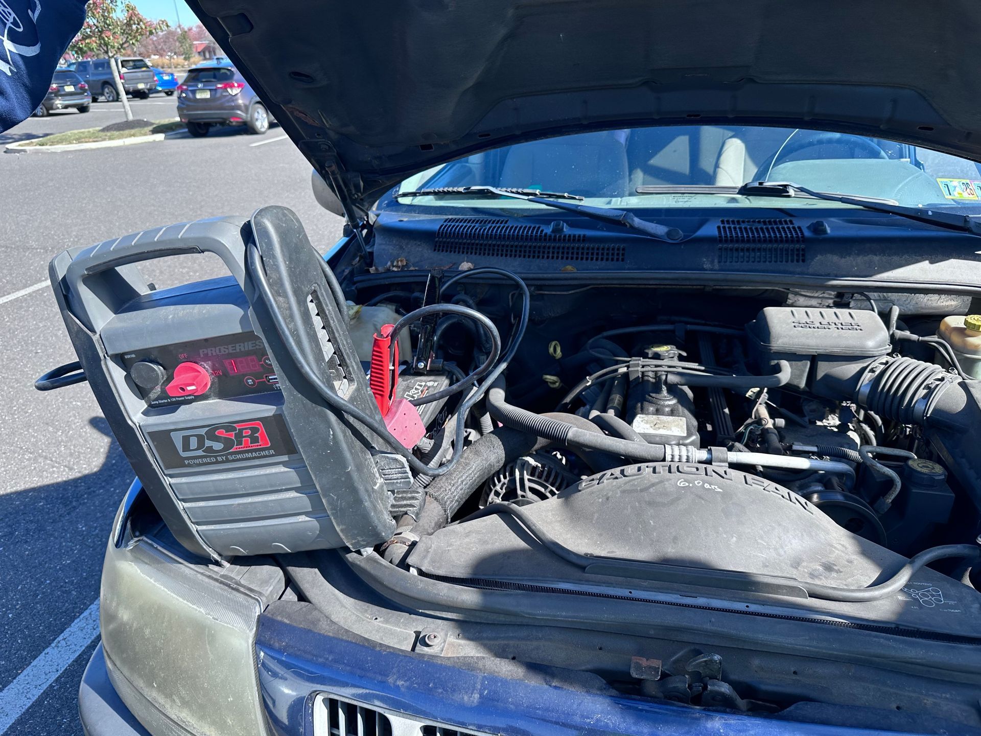 Car engine bay with hood open and a battery jump starter clipped to the battery.