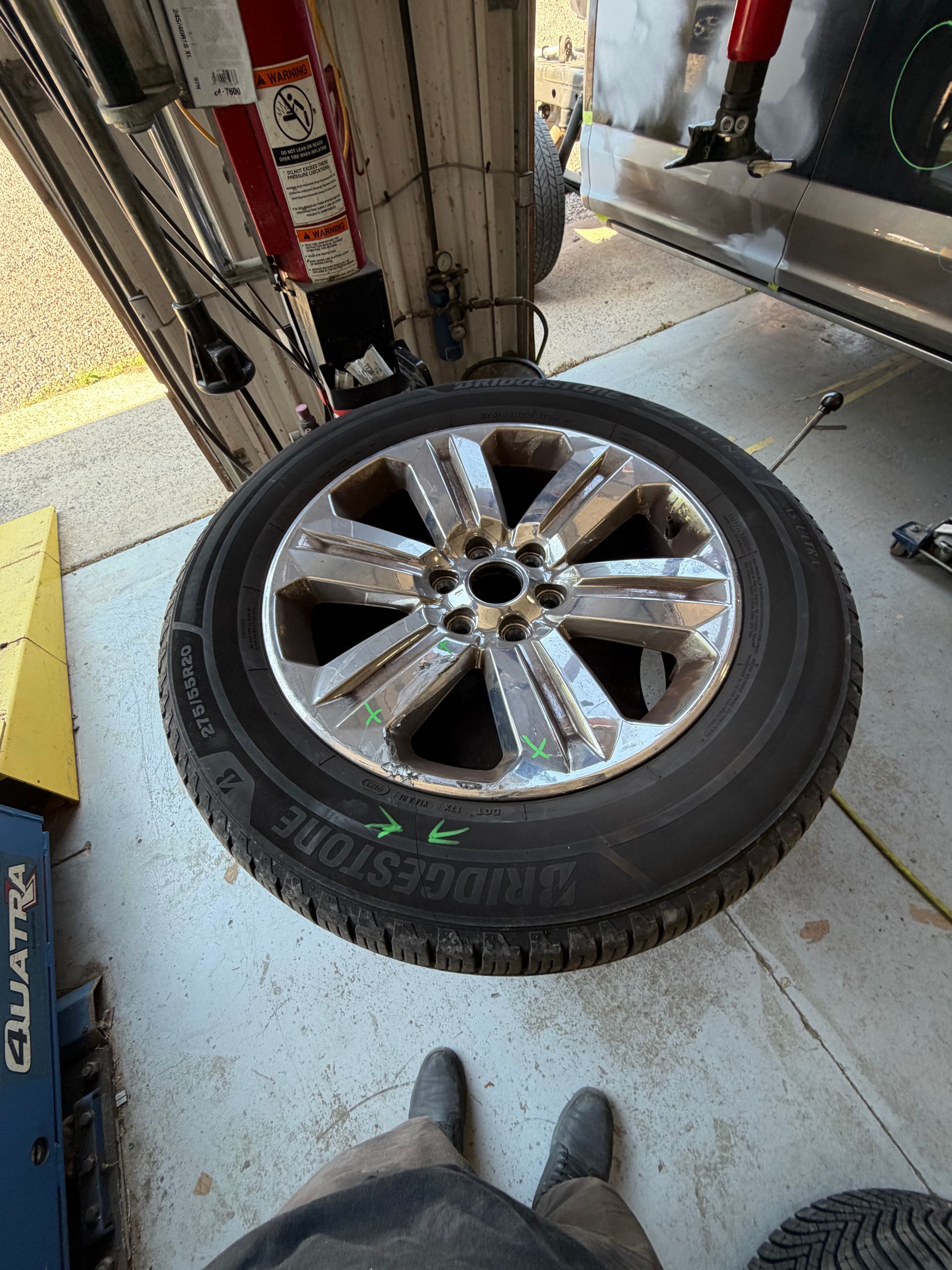 Car tire with chrome rim on a garage floor beneath a vehicle lift, viewed from above
