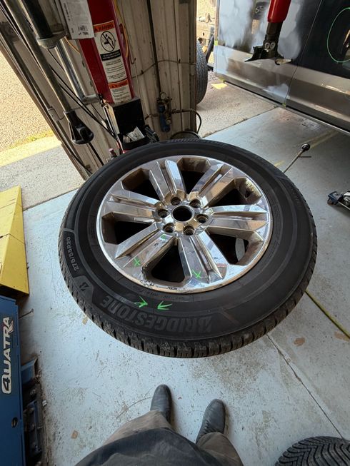 Car wheel and tire on the floor beside a lift in an auto shop, with a person’s shoes visible at the bottom.