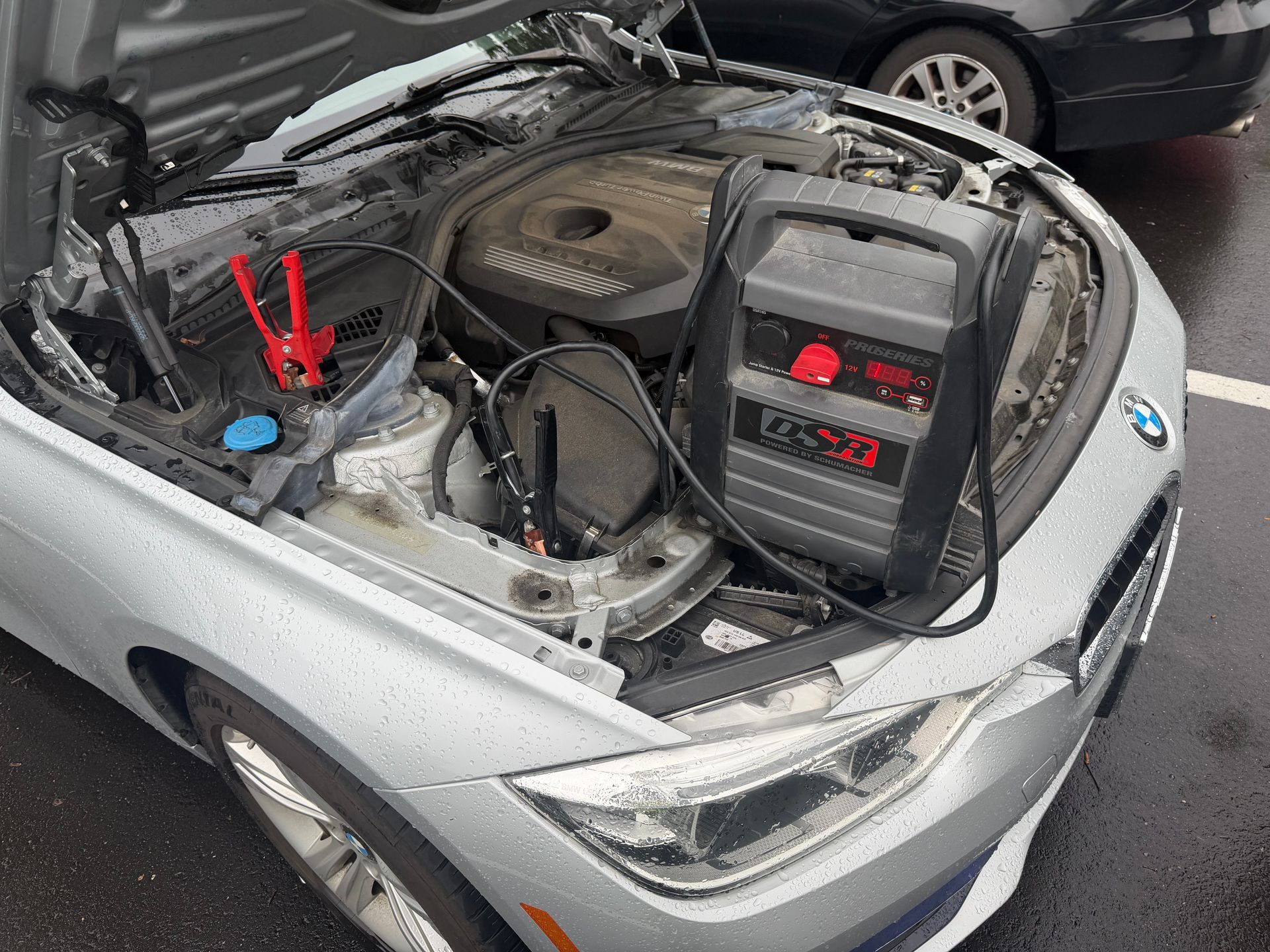 Silver car with hood open, showing engine bay and a jump starter connected to the battery.