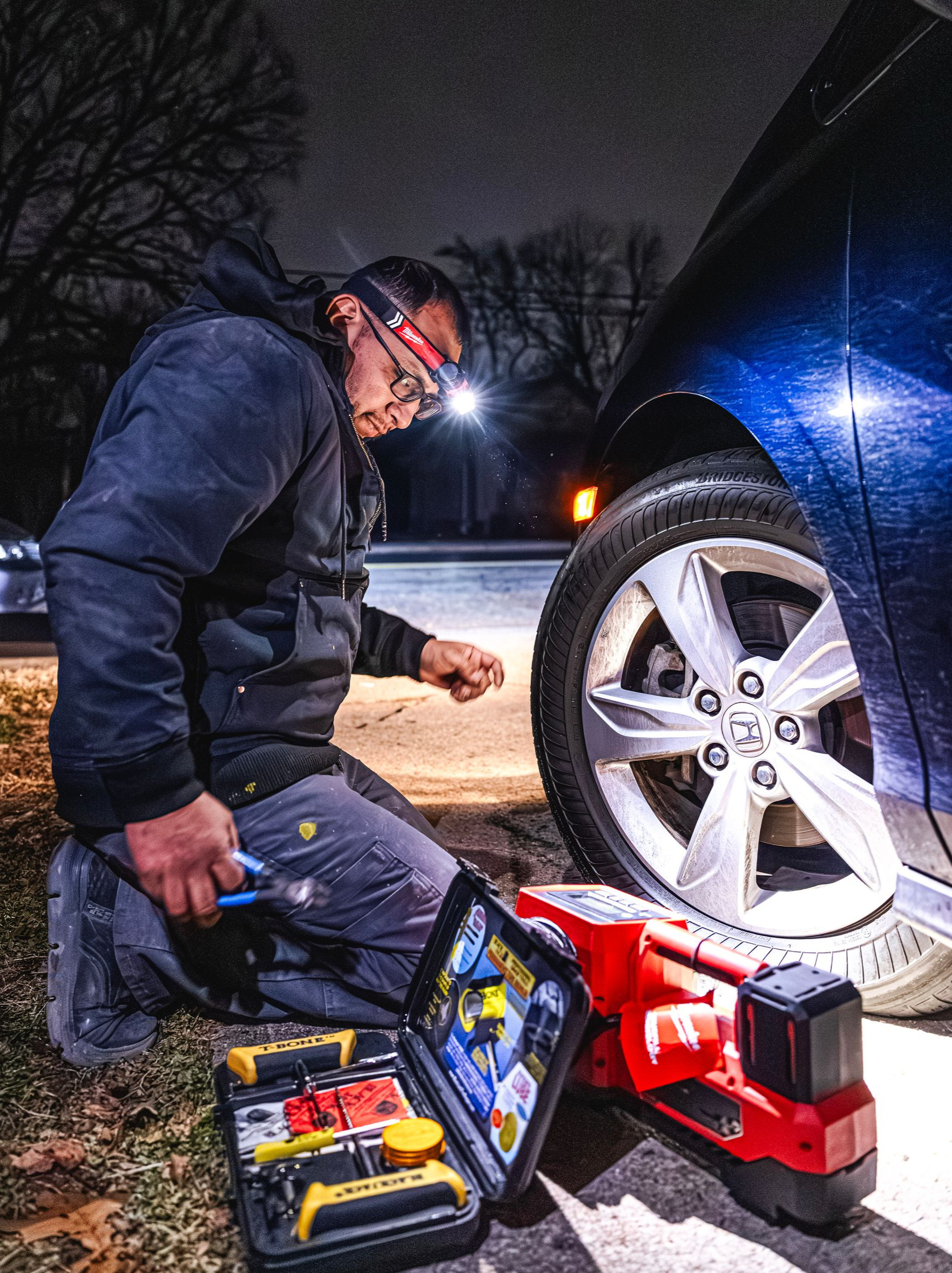 Mechanic kneeling beside a car wheel at night, using a red jack and tool kit to change a tire.