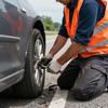 Roadside worker in orange vest kneeling to change a car tire on the shoulder