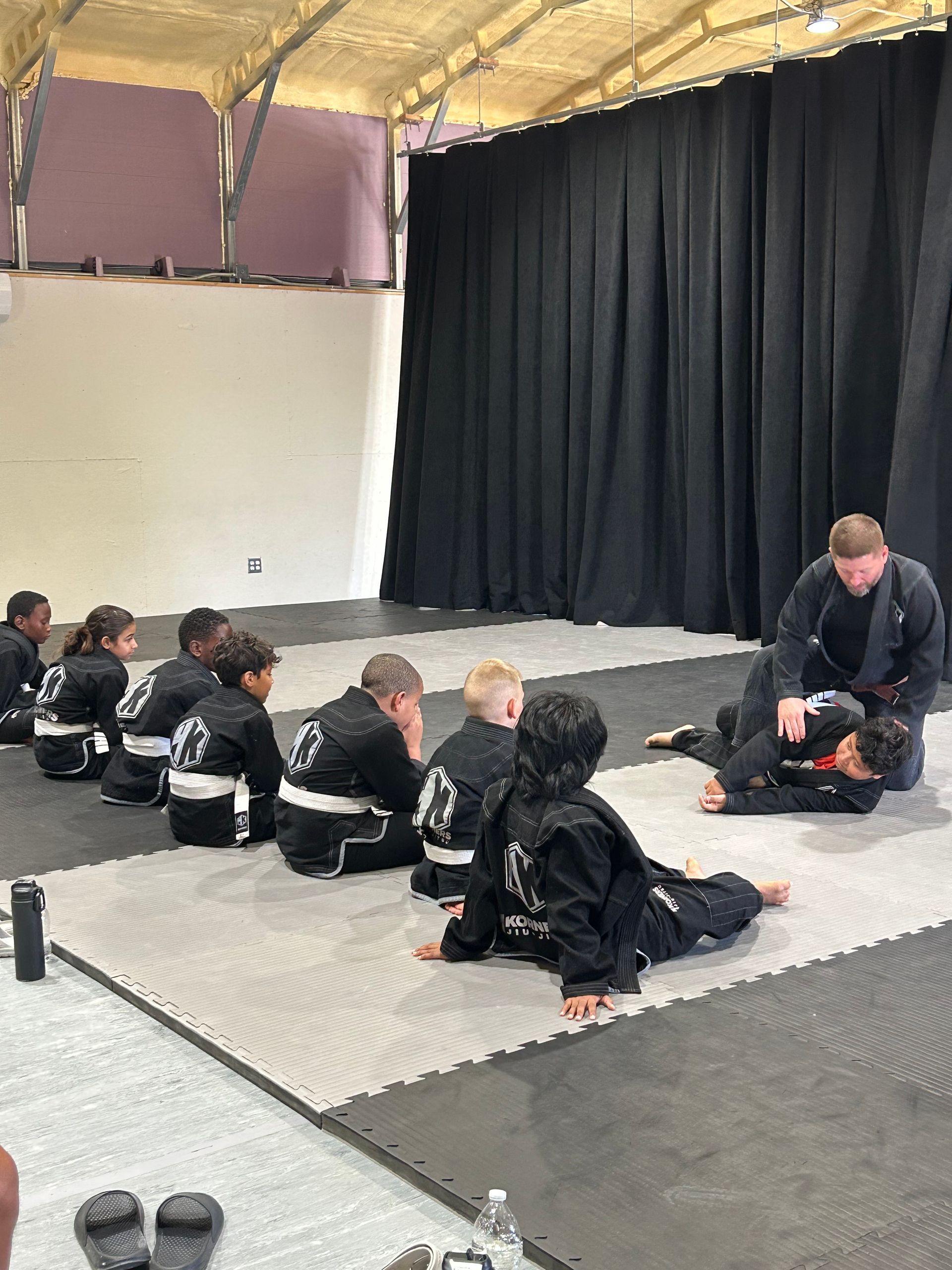 Children seated on mats in 4Korners Jiu-Jitsu in Princeton, TX, watching an instructor on a stage with black curtains.