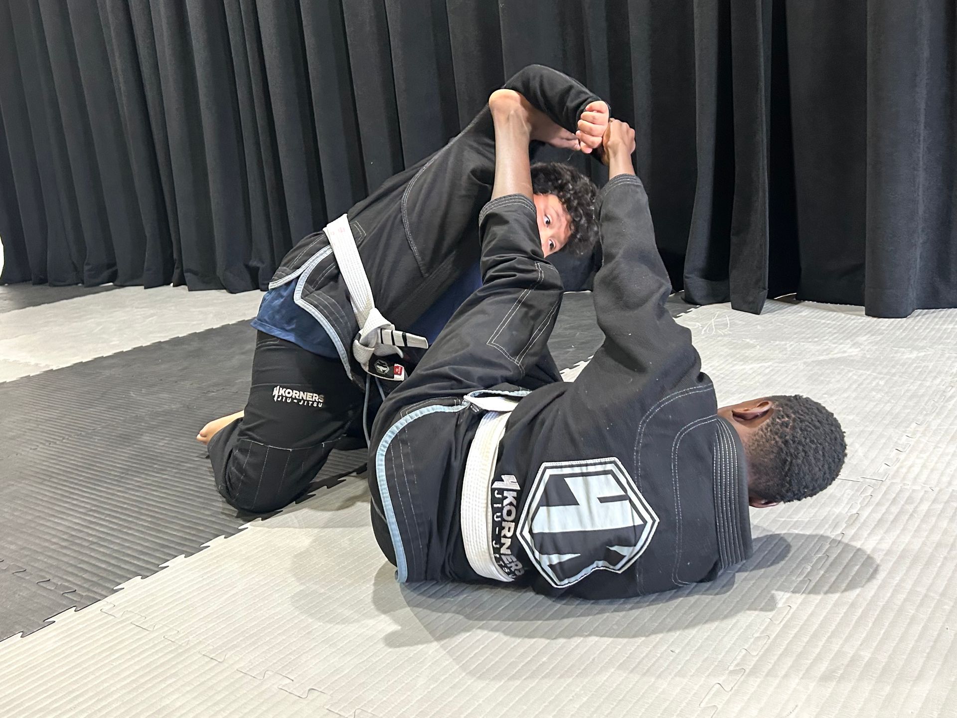 Martial arts class with people stretching on a mat, led by a seated instructor in a black gi at 4Korners Jiu-Jitsu in Princeton, TX