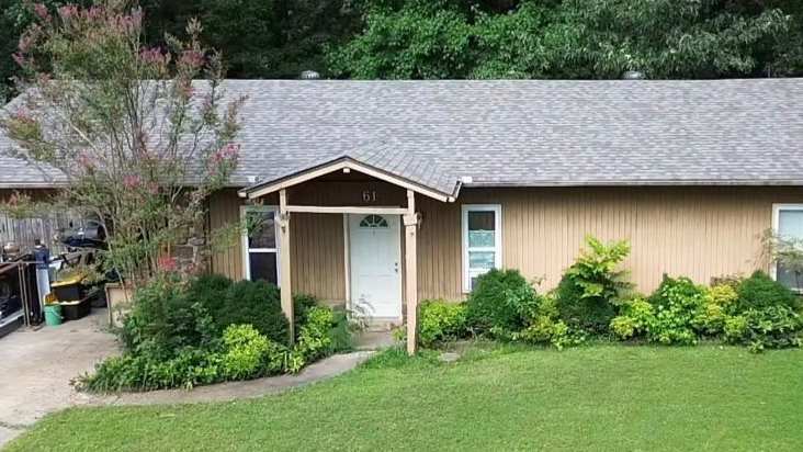 A single-story tan house with a gray shingled roof, front porch, and surrounding green landscaping on a sunny day.