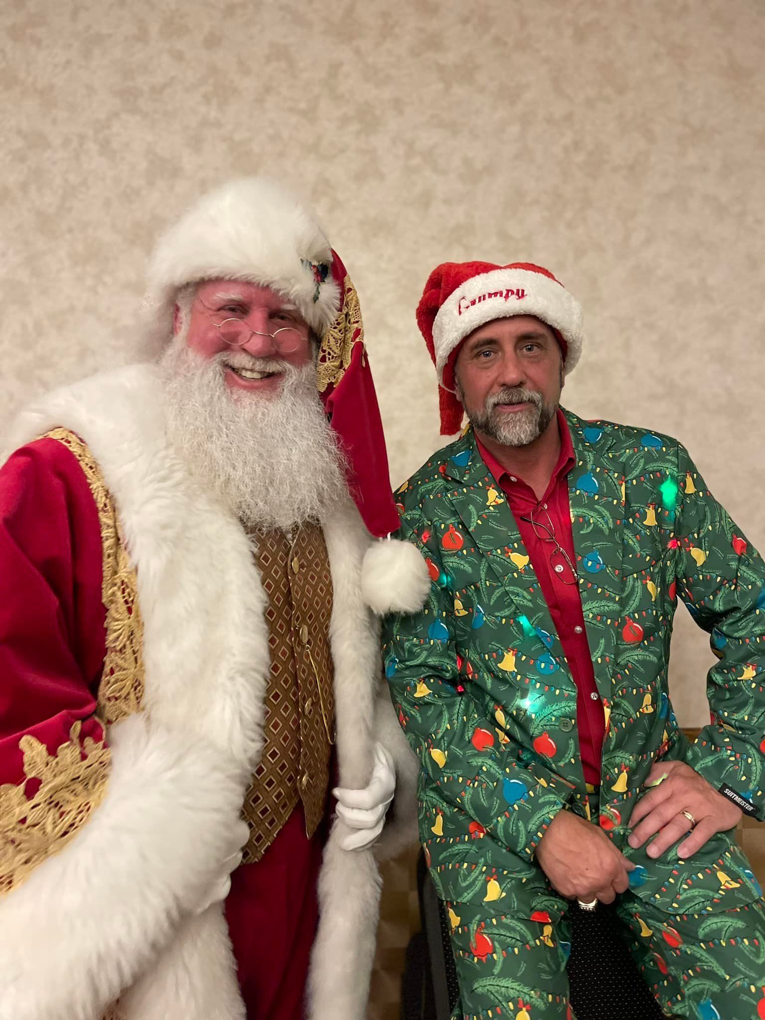 Santa Claus standing next to a man wearing a green holiday-patterned suit and a matching Santa hat against a plain wall.