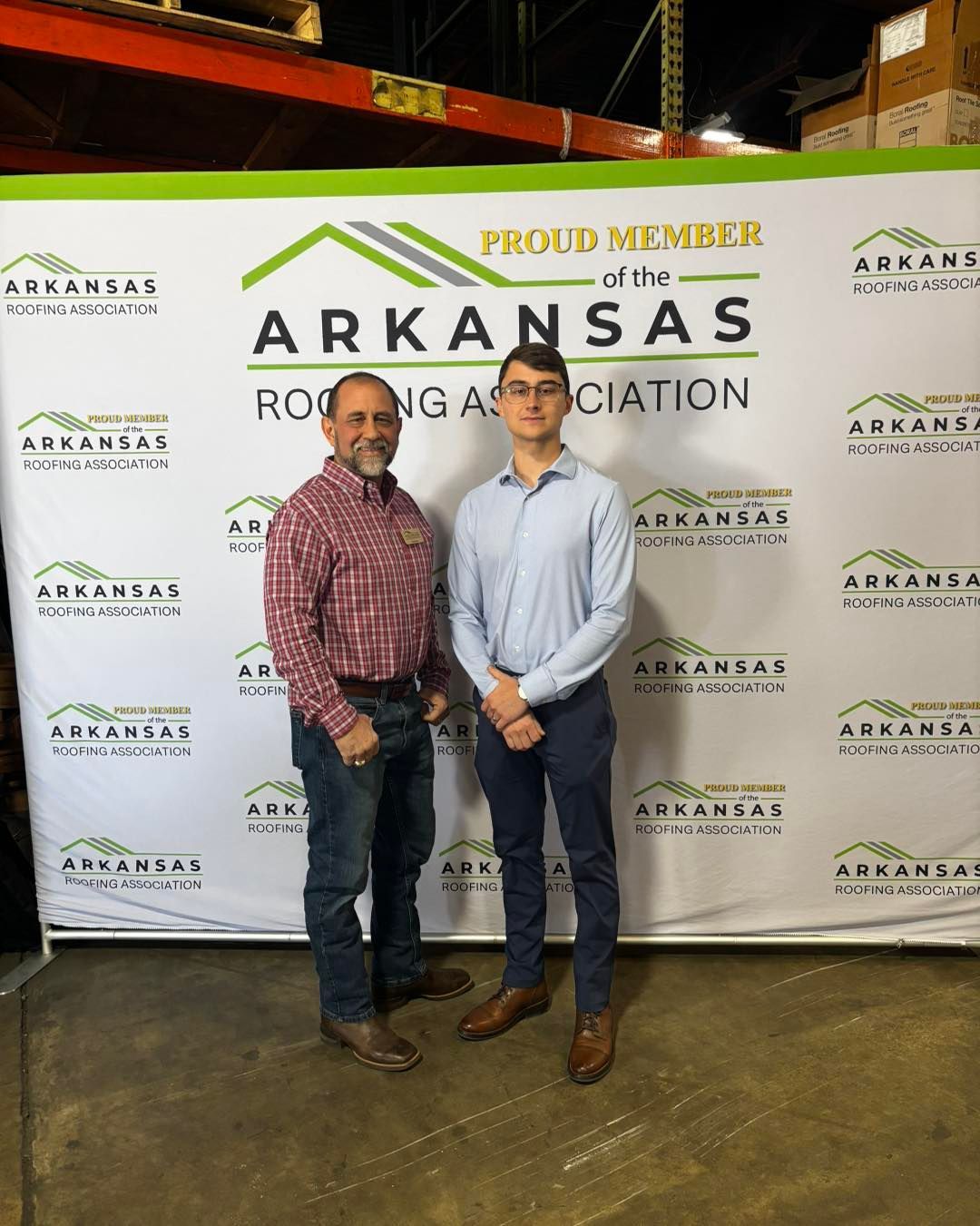 Two professionals stand smiling in front of an Arkansas Roofing Association branded backdrop in a warehouse setting.