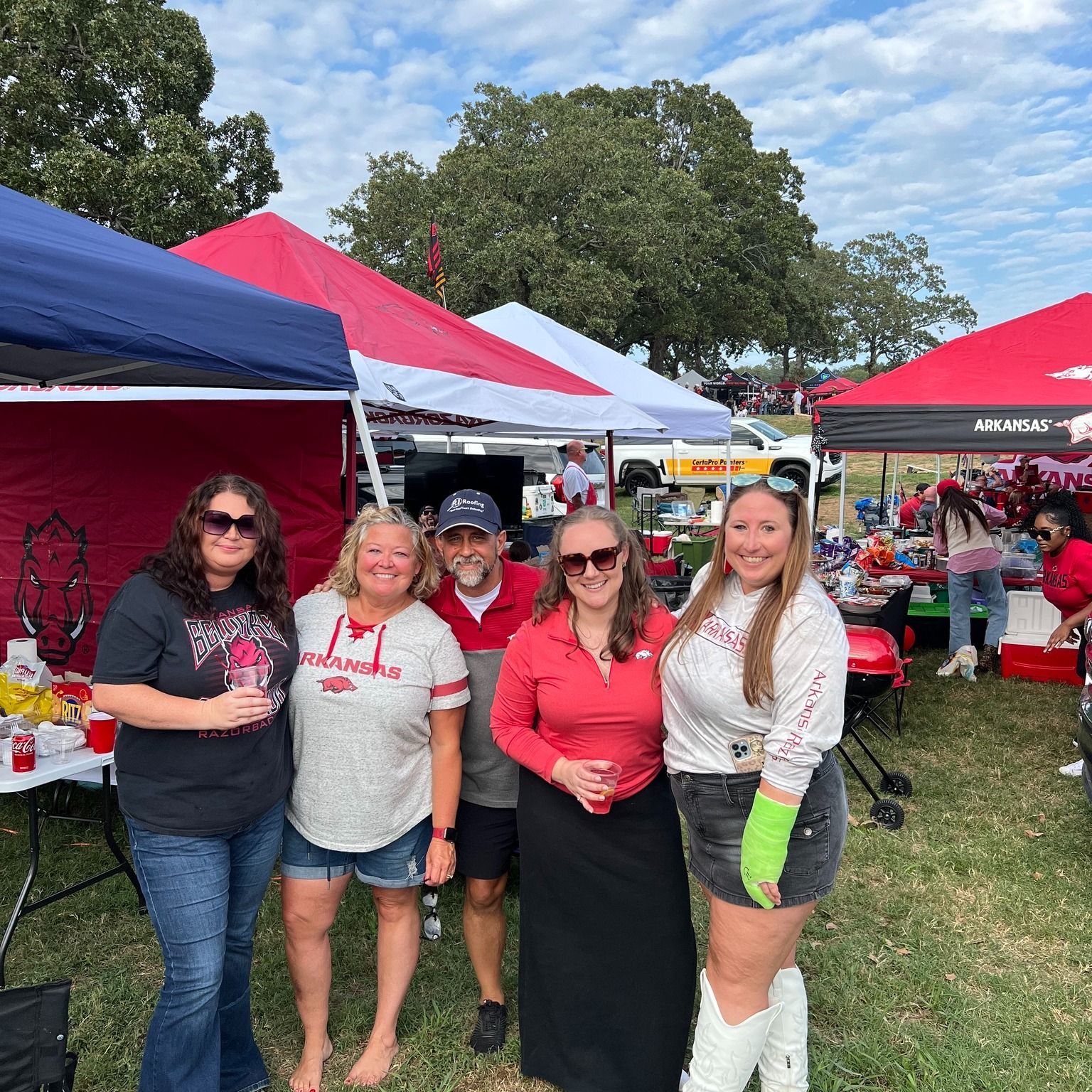 Four people pose smiling under red and white tailgating tents on a sunny day, wearing casual team-themed apparel.