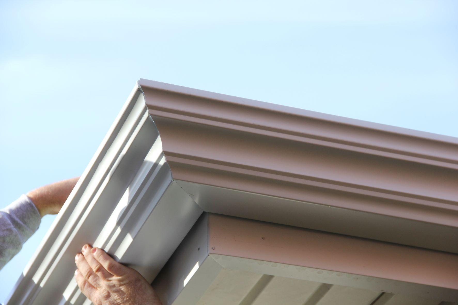 Hands installing a white decorative crown molding trim piece onto the corner of a house exterior against a blue sky.