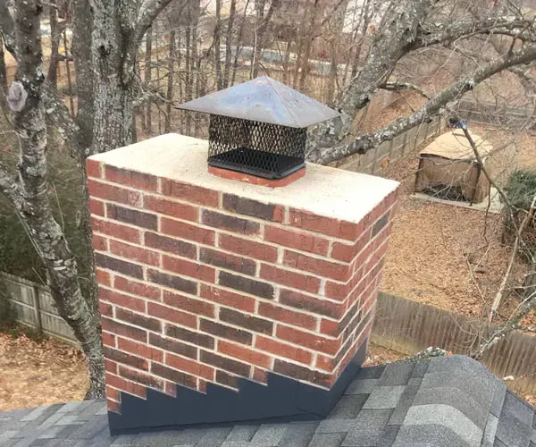 A brick chimney with a metal cap and mesh guard installed on a shingled roof.