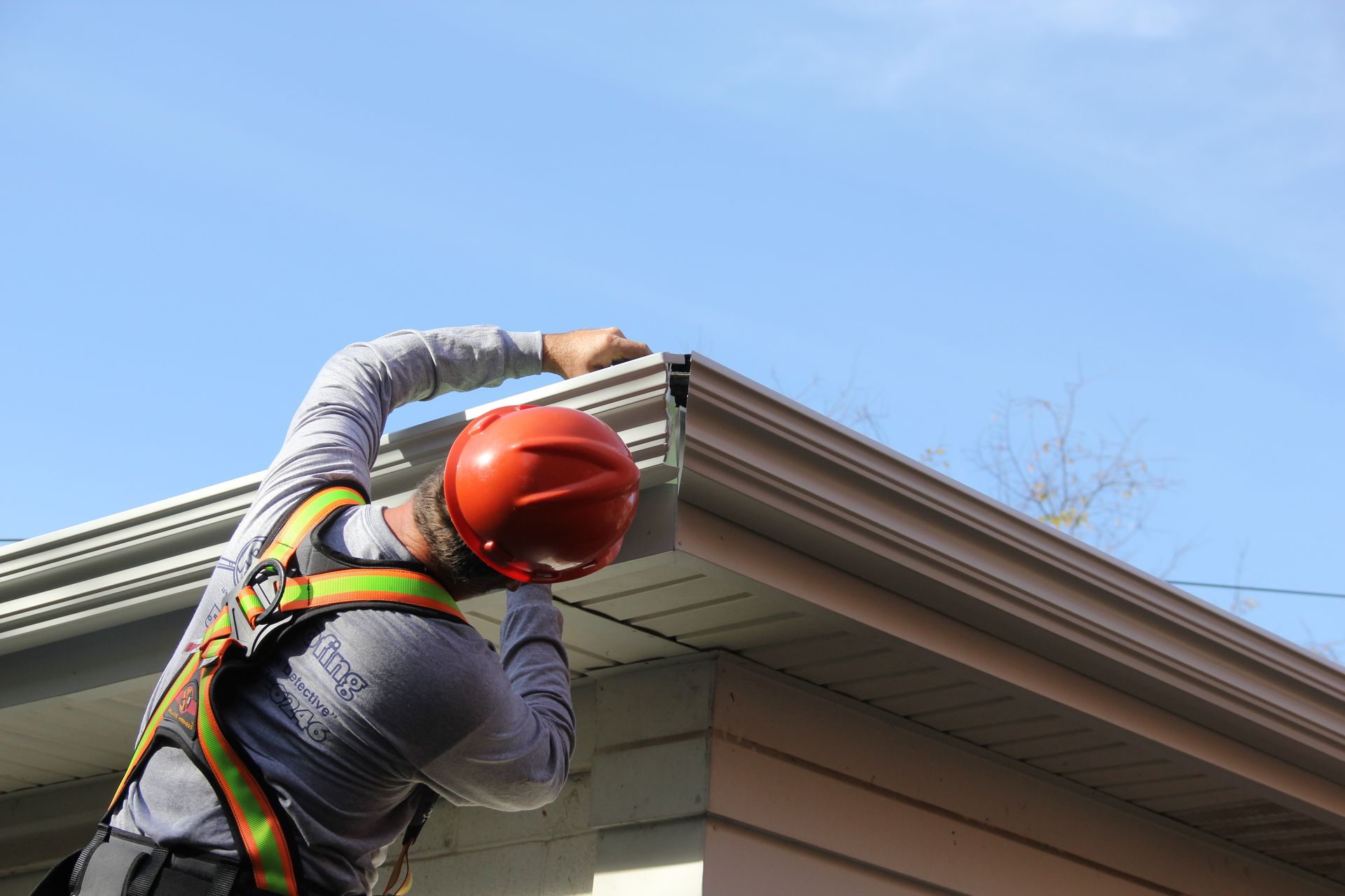A worker in a safety harness and orange hard hat installs or repairs a light-colored gutter on the edge of a roof.