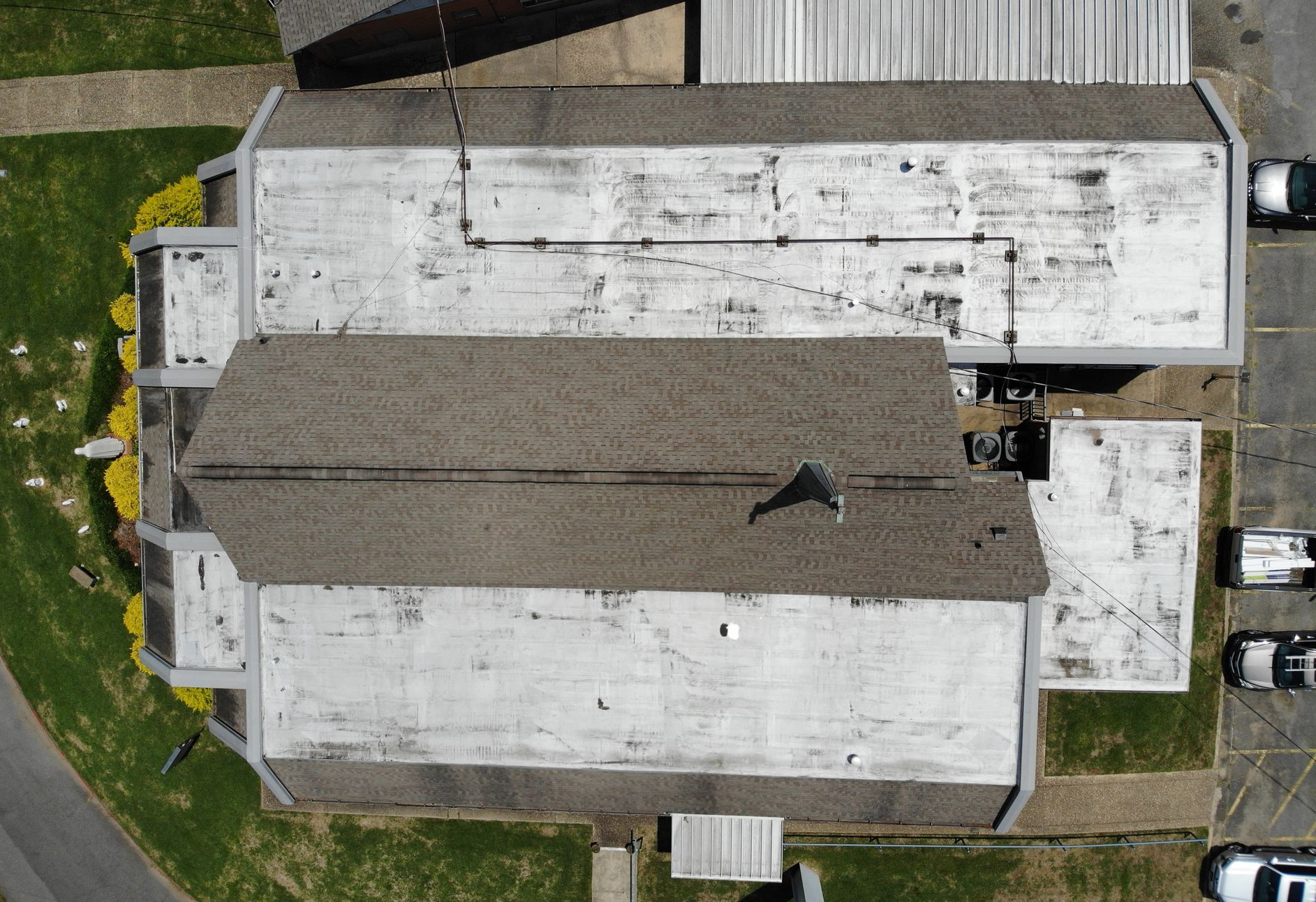 A high-angle aerial view of a building roof, showing a central shingled section flanked by flat, white, weathered sections.