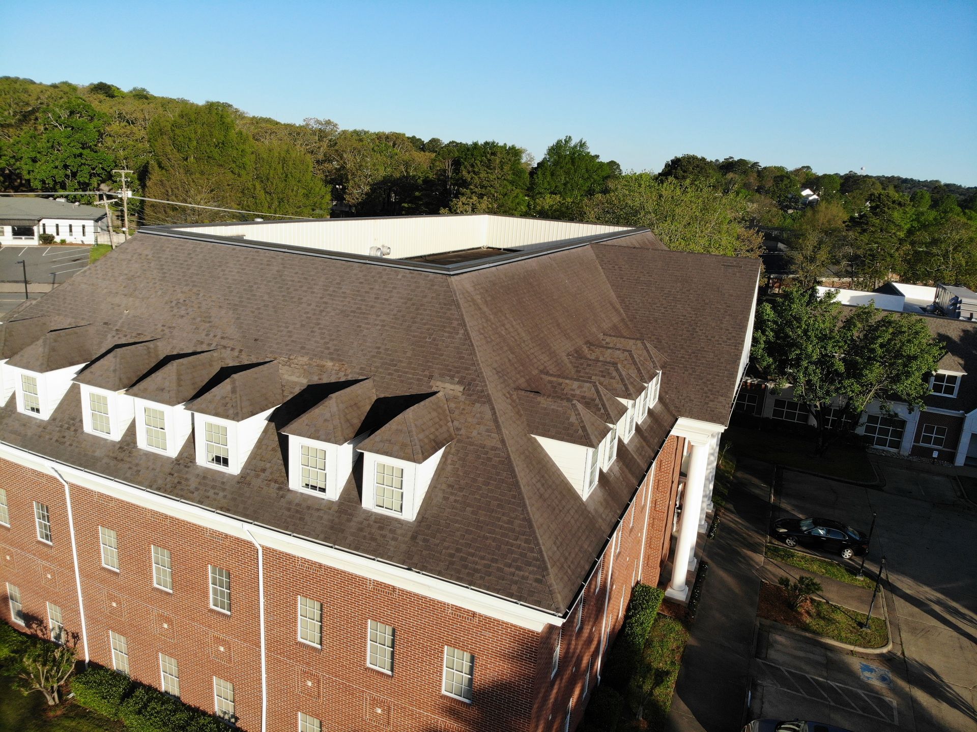 High-angle shot of a brick building with a dark shingled, multi-dormer roof and a central flat rooftop section.