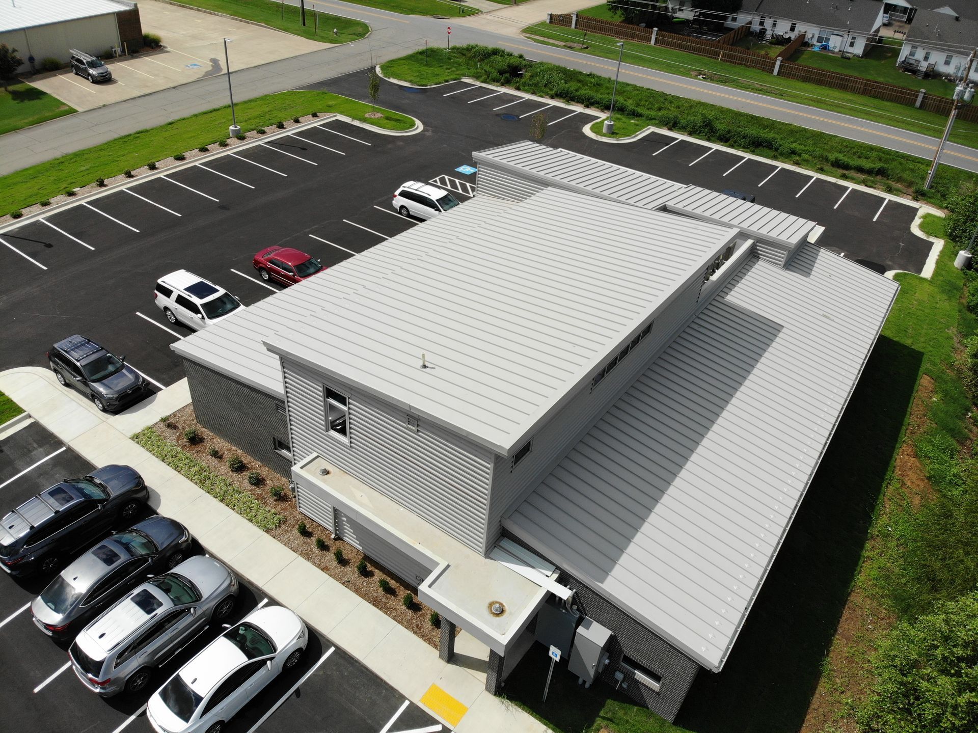 Aerial view of a gray, modern commercial building with a metal roof and a parking lot with several parked cars.