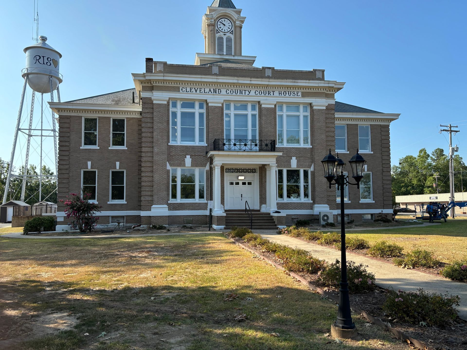 A brick courthouse with a clock tower and a large, elevated white water tower on the left side under a clear blue sky.