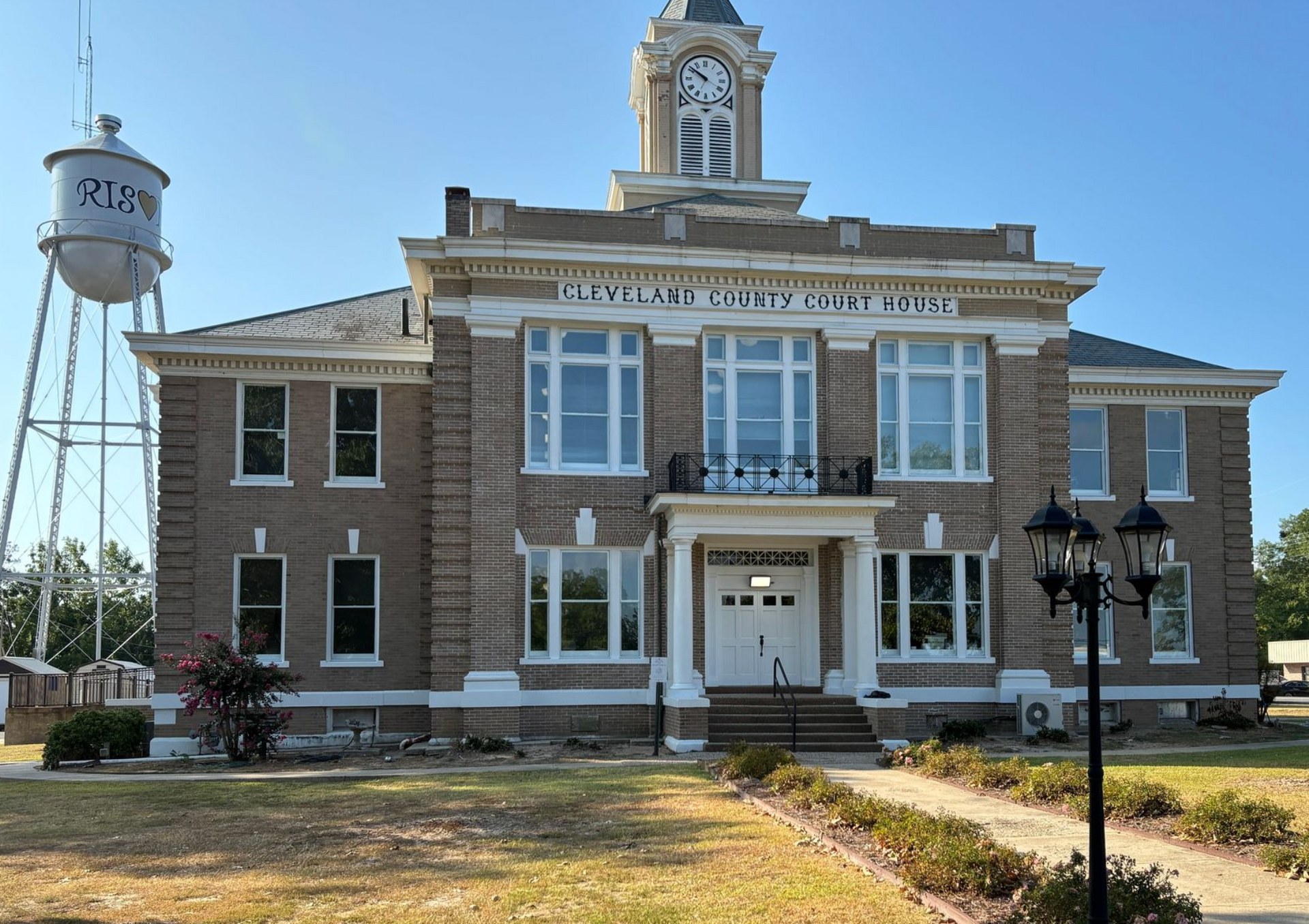 A brown brick, two-story Cleveland County Courthouse with a central clock tower, next to a water tower under a blue sky.