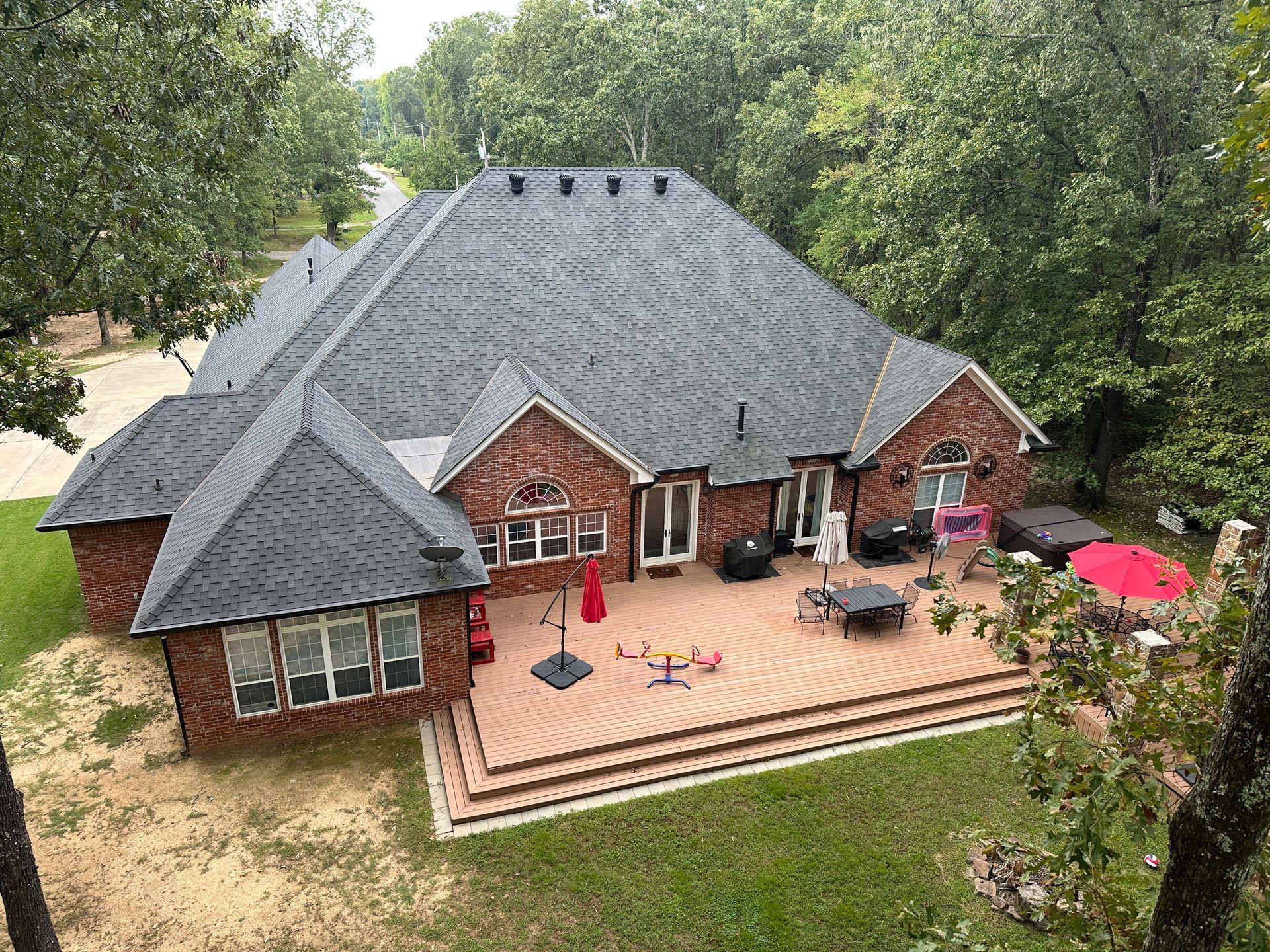 High-angle view of a brick house with a large composite deck, backyard patio furniture, and surrounding woods.