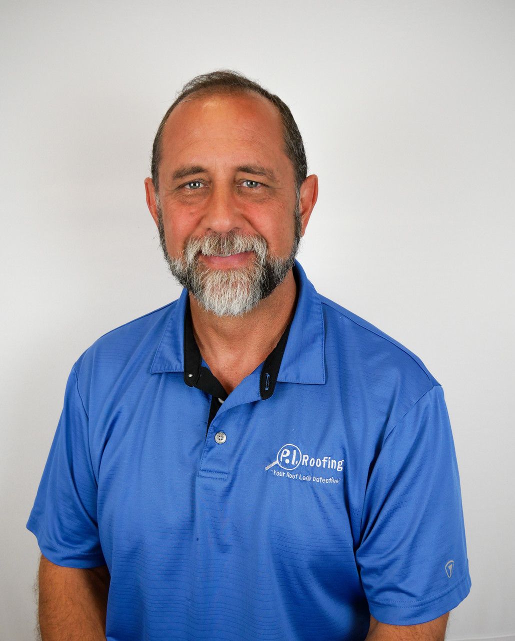 A smiling person with a grey beard and short hair, wearing a blue polo shirt with a white logo, against a white background.
