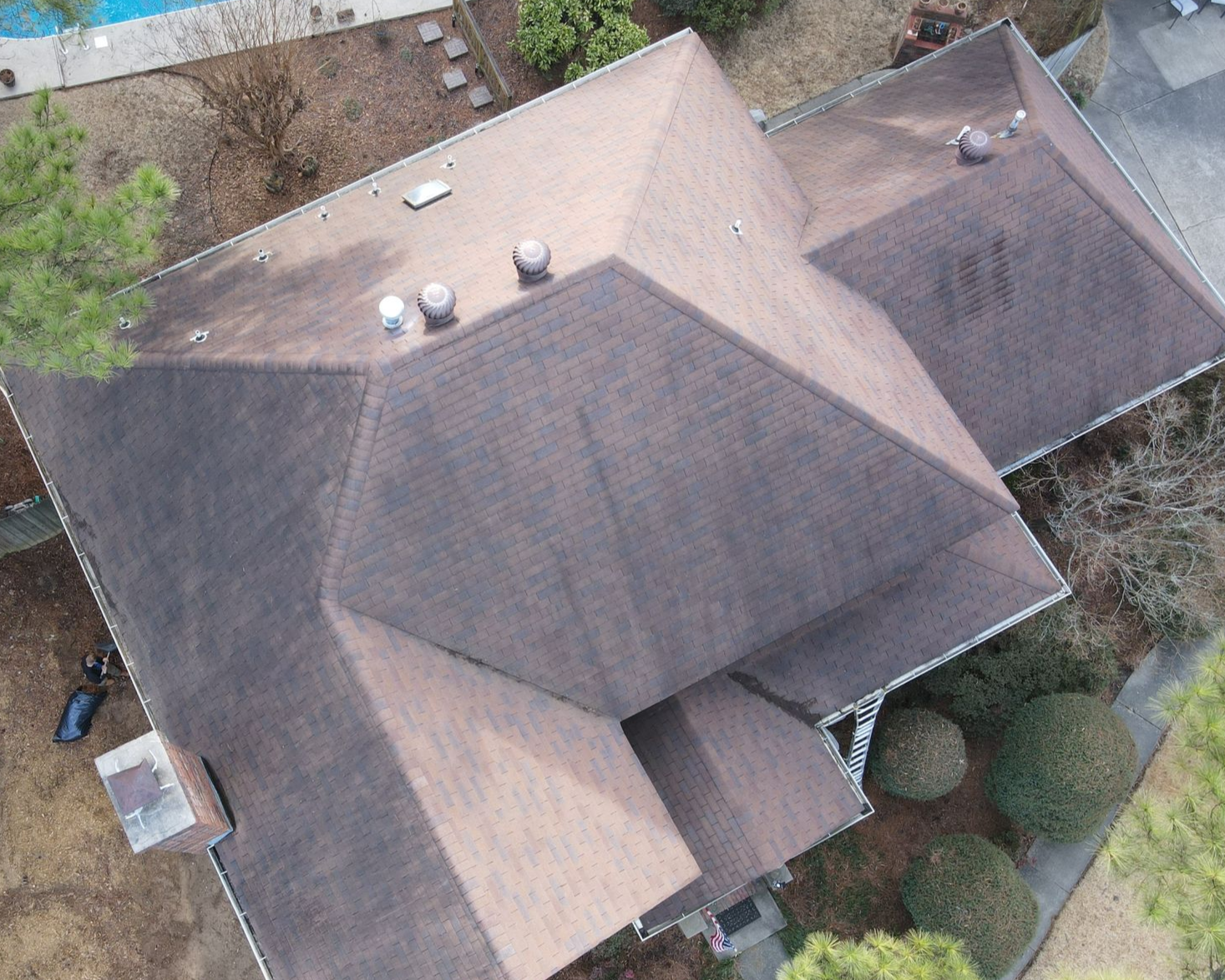 An aerial view of a dark shingled residential roof with multiple peaks, chimneys, and vents, surrounded by trees.