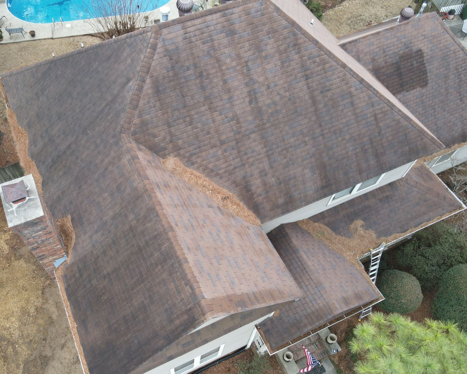 An aerial view of a brown asphalt-shingled roof on a multi-level house, with scattered fallen leaves in the roof valleys.