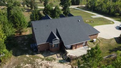 An aerial view of a brick house with a dark shingled roof, a wraparound deck, and a driveway set in a rural landscape.