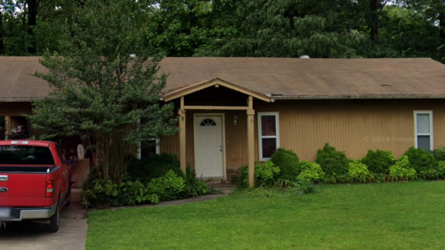 A single-story, tan-colored house with a brown roof and a small front porch, with a red truck parked in the driveway.