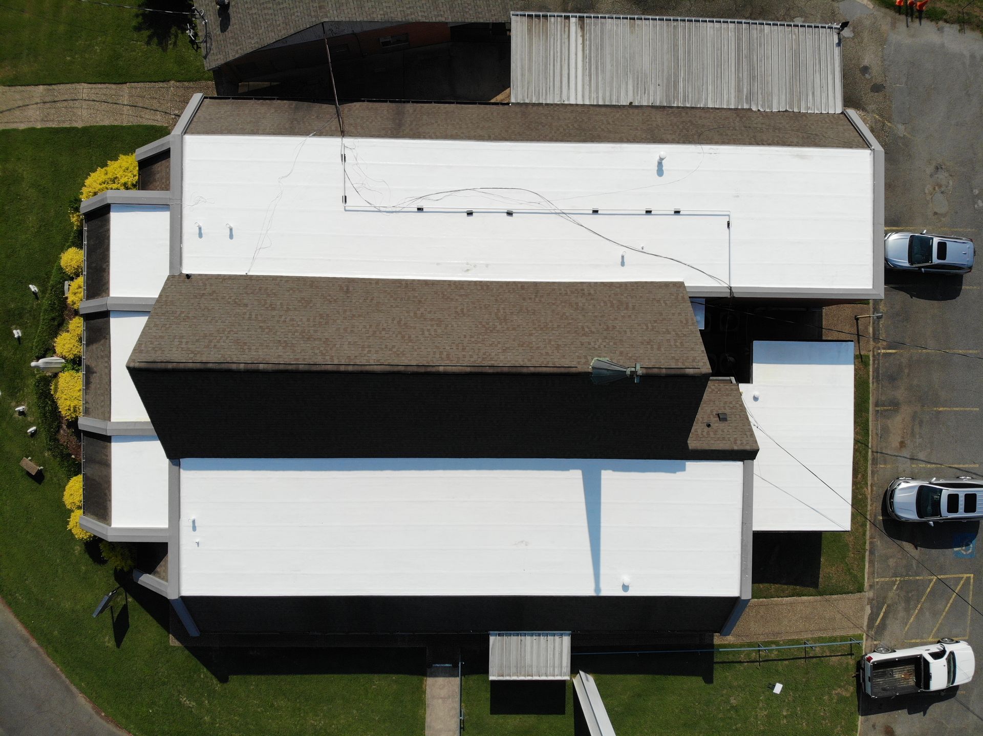 An aerial view of a building with a multi-section roof, featuring a brown shingled peak and white flat roof sections.