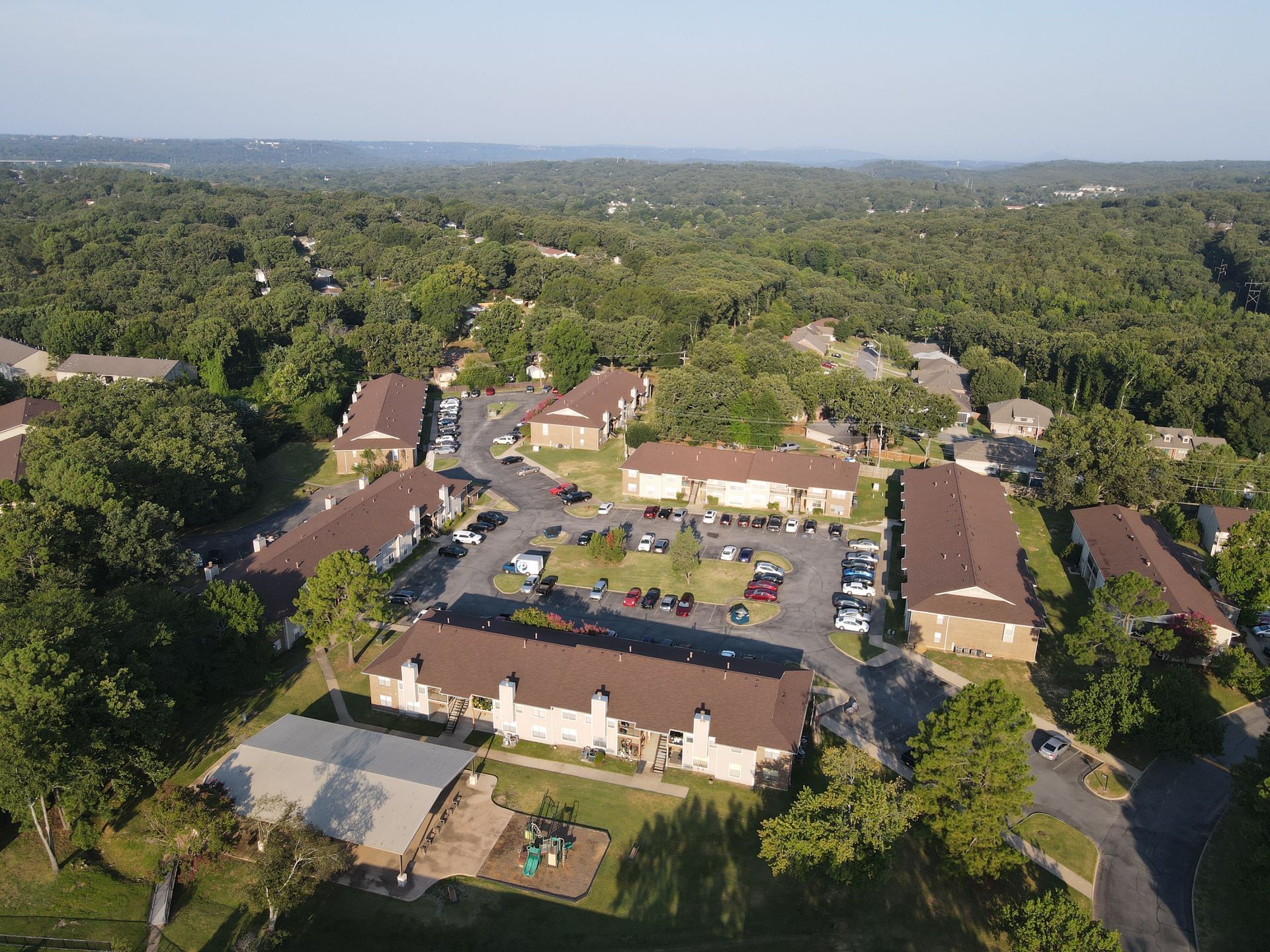 Aerial view of a residential apartment complex with tan buildings, dark roofs, and surrounding wooded landscape.