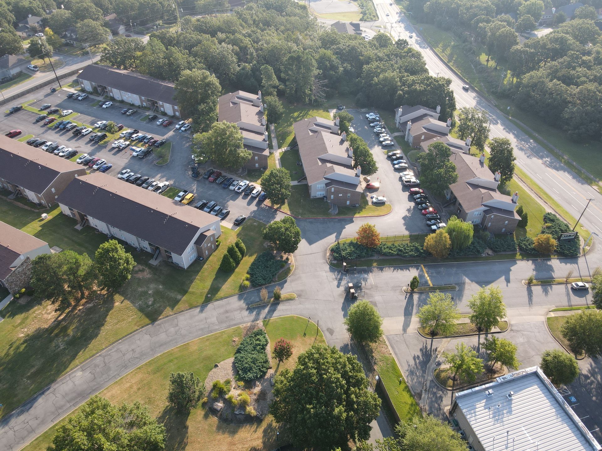 Aerial view of an apartment complex with several buildings, parking lots, and green spaces surrounded by trees.