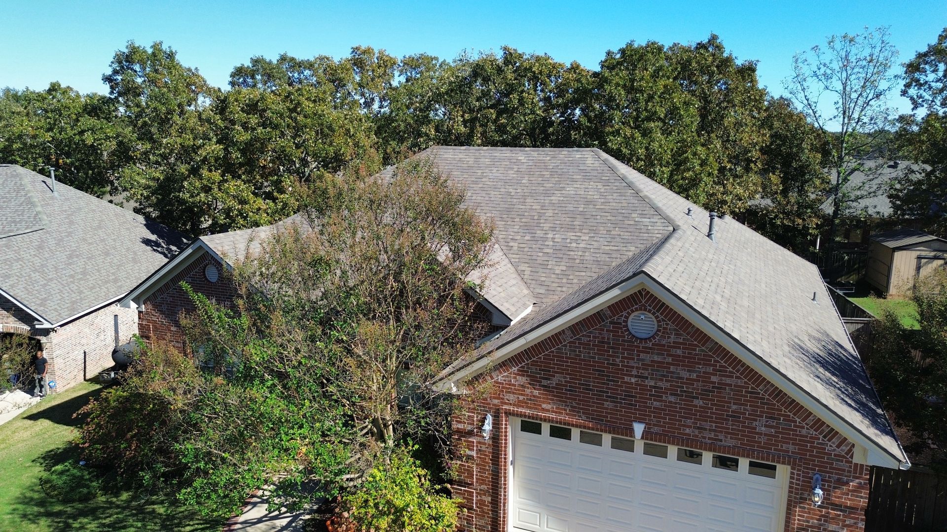 Aerial view of a brick house with a multi-pitched gray shingle roof, surrounded by trees under a clear blue sky.