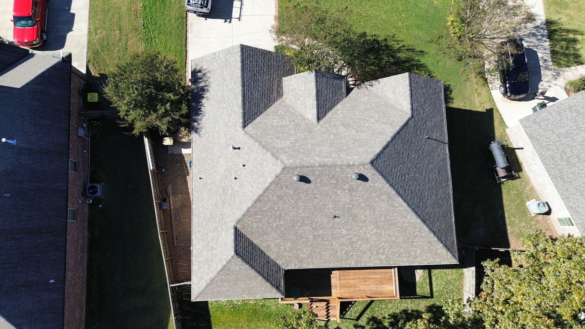 Aerial view of a residential house with a gray shingled roof, a wooden deck, and surrounding lawns and driveways.