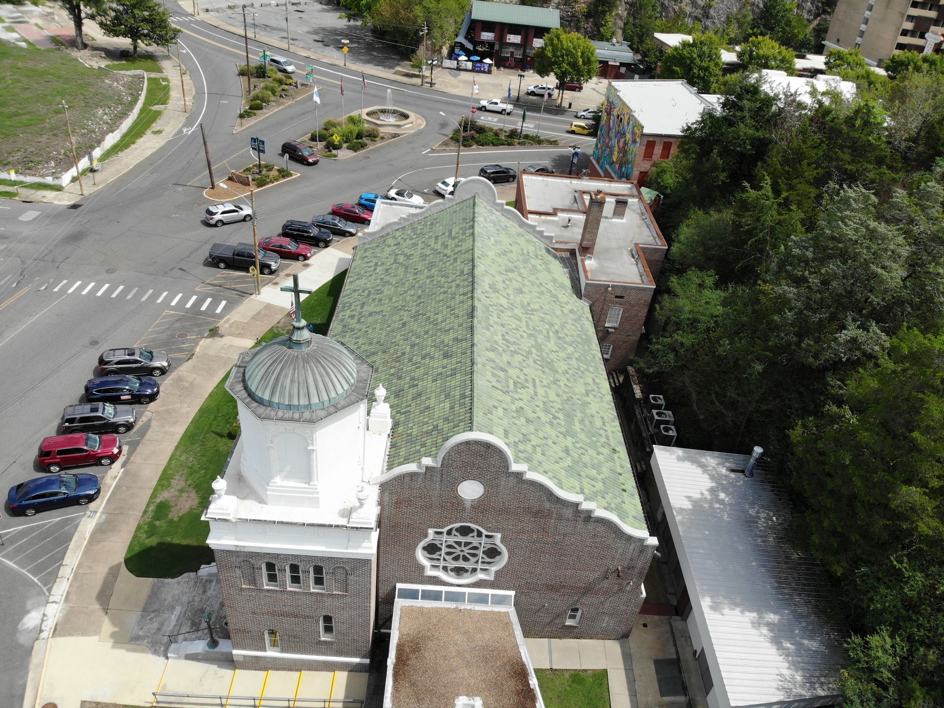 Aerial view of a church with a green tiled roof and white domed tower, situated next to a parking lot and street.